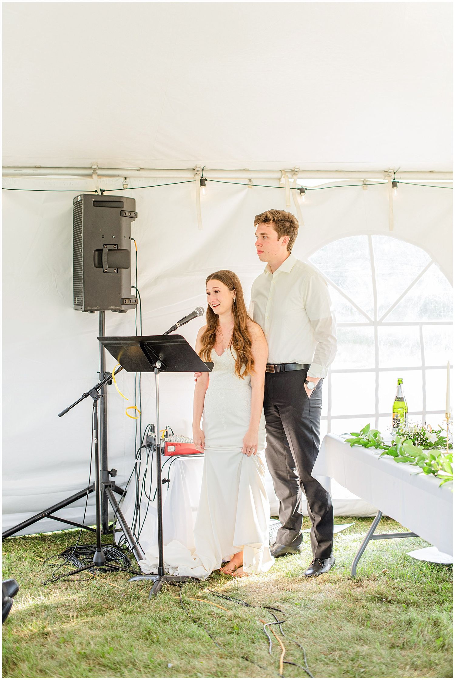 Bride and groom stand together at a microphone during their outdoor wedding reception under a white tent.