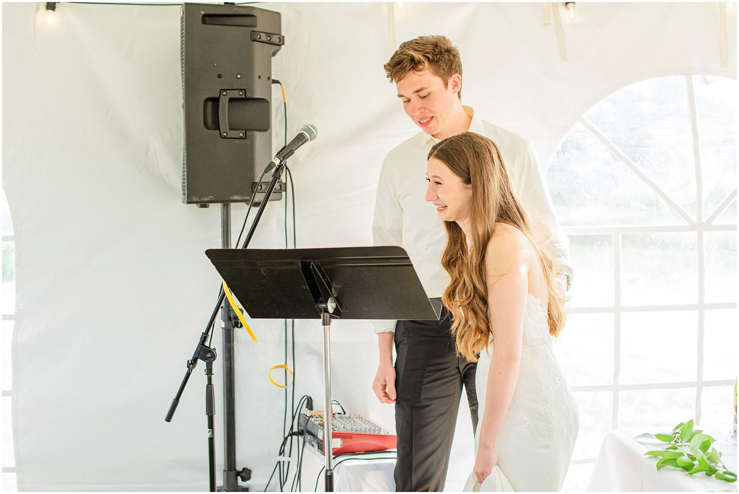 Young couple reviewing sheet music at microphone stand during outdoor wedding reception under white tent.