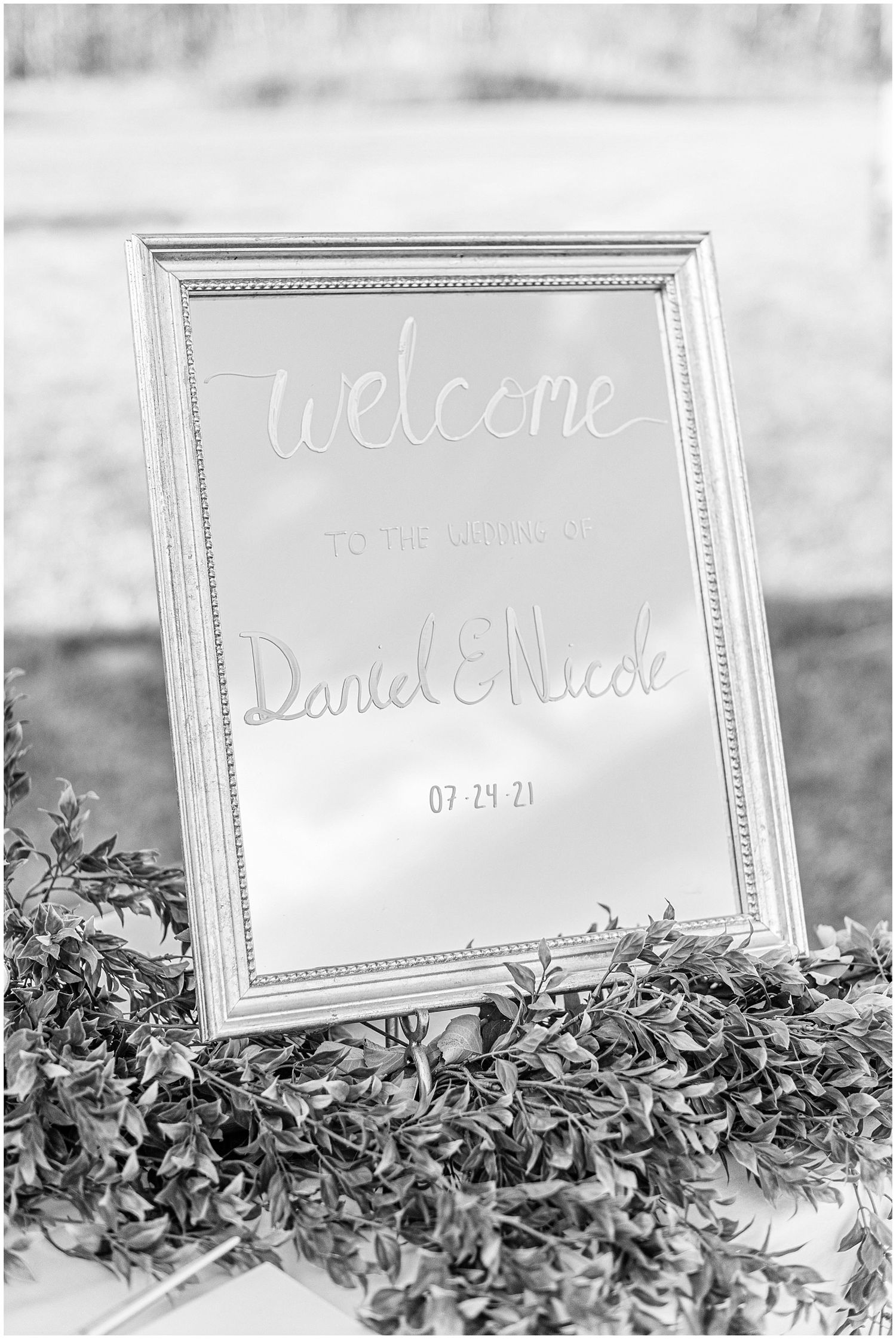 Black and white welcome mirror sign for Daniel and Nicole wedding dated 07.24.21 surrounded by green foliage.