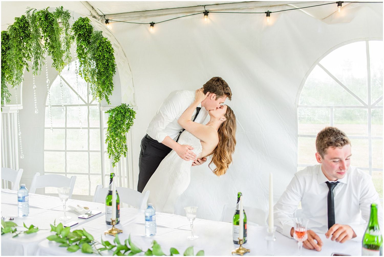 Groom dips bride for a kiss at elegantly decorated wedding reception table under white tent.