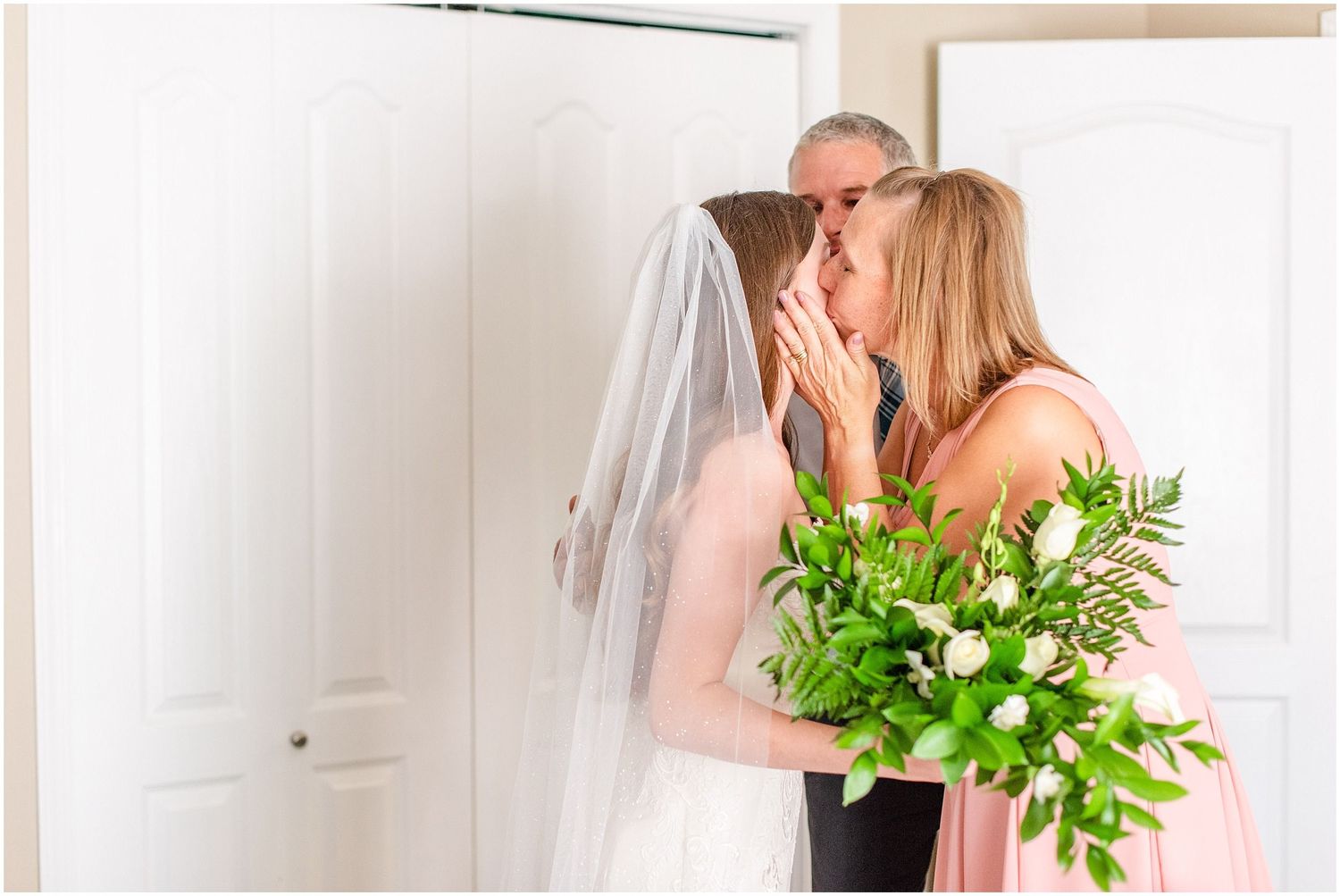 Bride in white gown and veil kissing her mother in pink dress while holding a green bouquet.