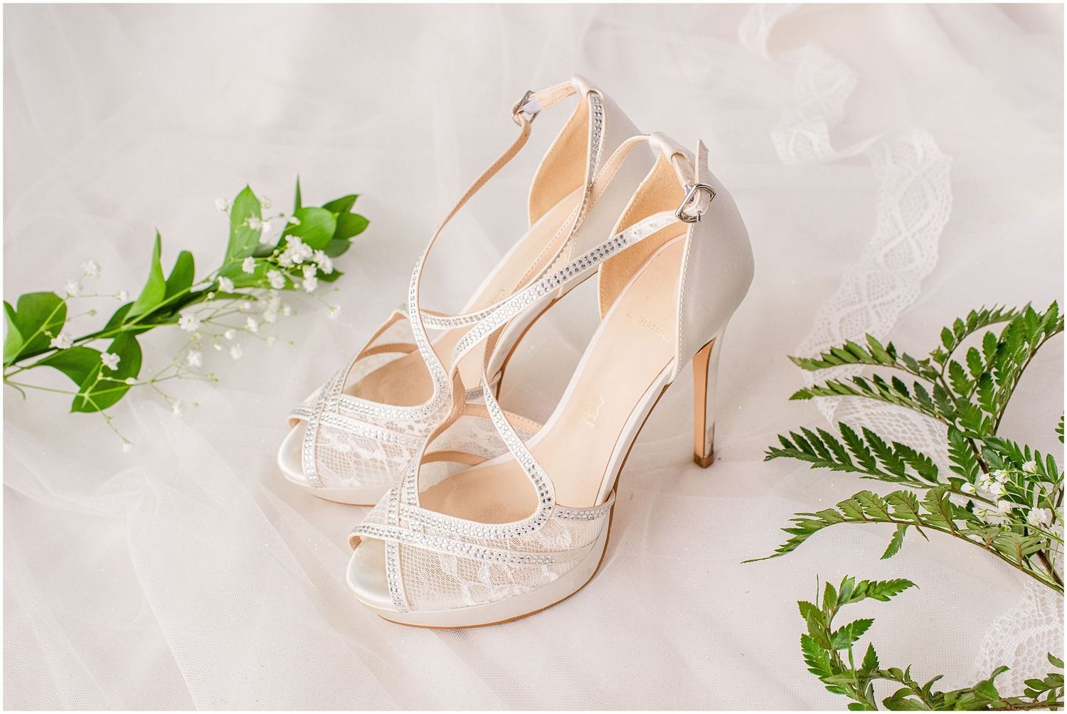 Elegant white lace bridal heels with crystal straps displayed on veil with greenery and baby's breath.