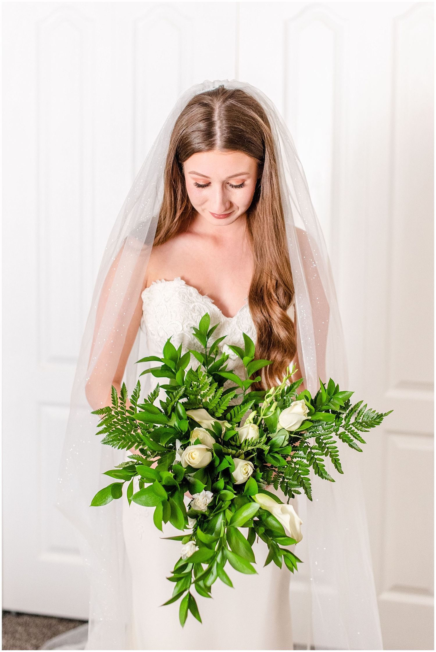 Bride in strapless lace gown and veil holding a lush green and white bouquet, looking down softly.