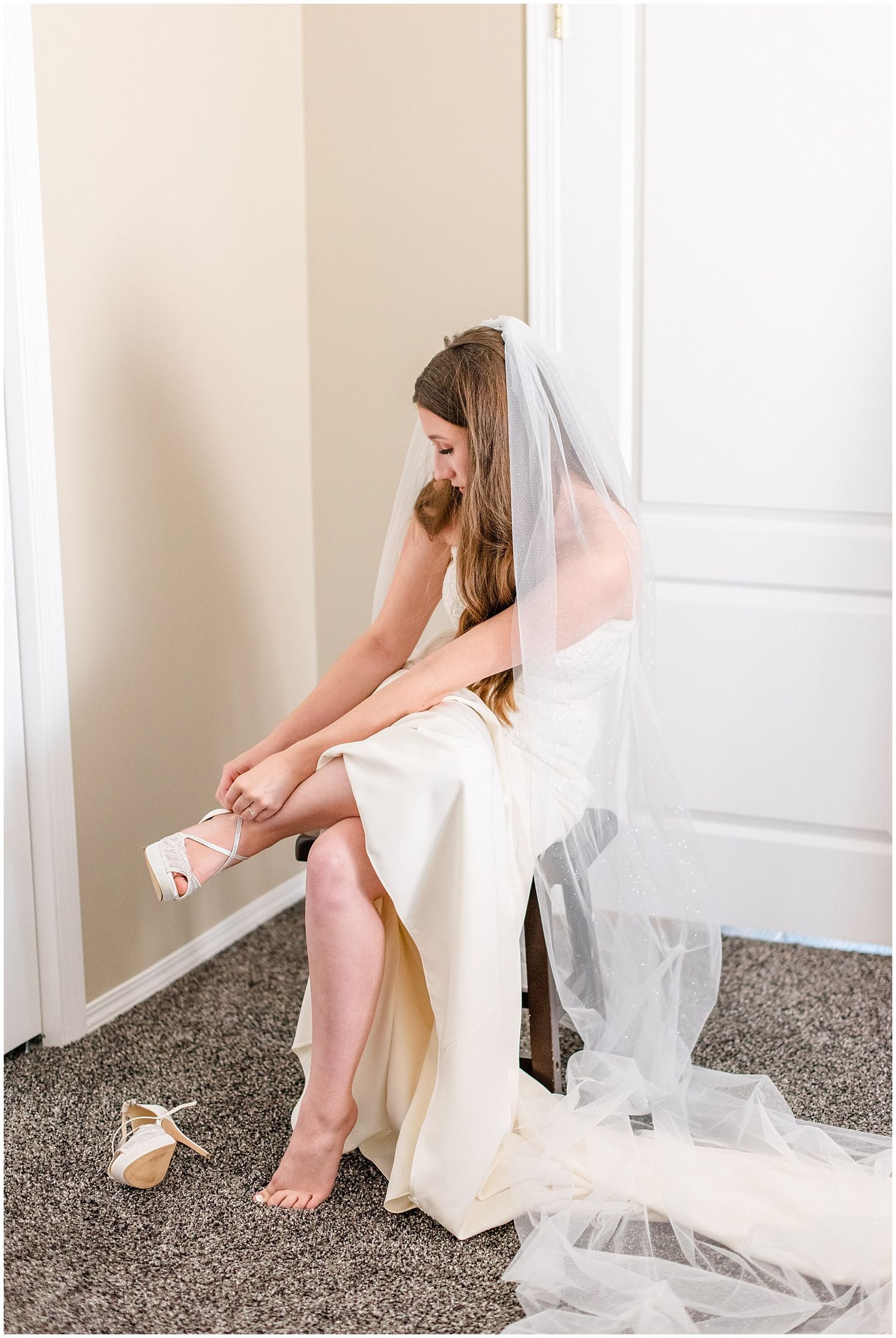 Bride in white gown and veil sitting and putting on lace heels in a softly lit room.