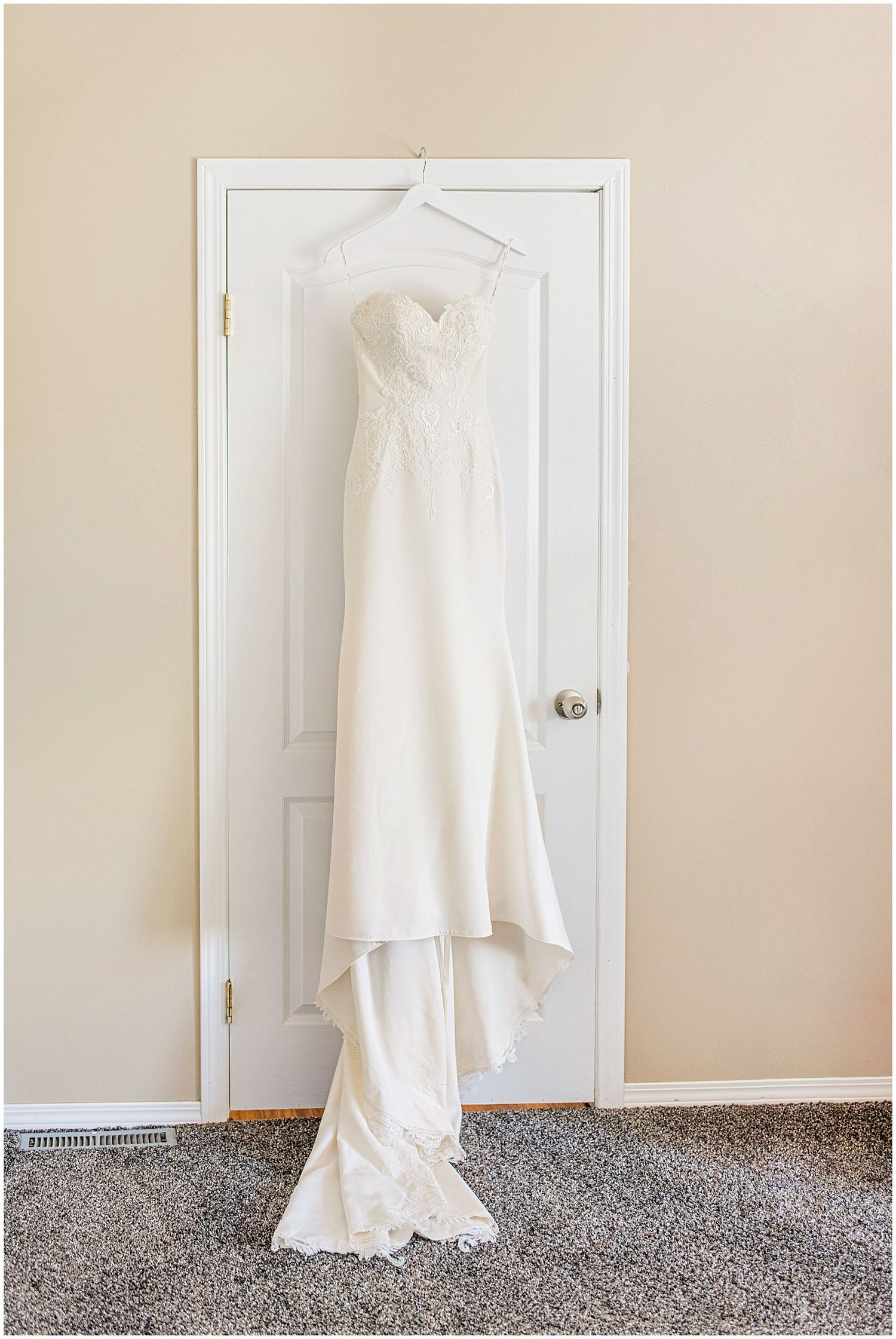 Elegant white strapless wedding gown with lace bodice hanging on a white door in a softly lit room.