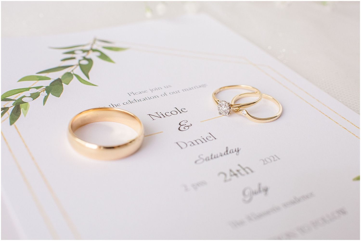 Gold wedding bands and diamond engagement ring displayed on elegant Nicole and Daniel wedding invitation with greenery.