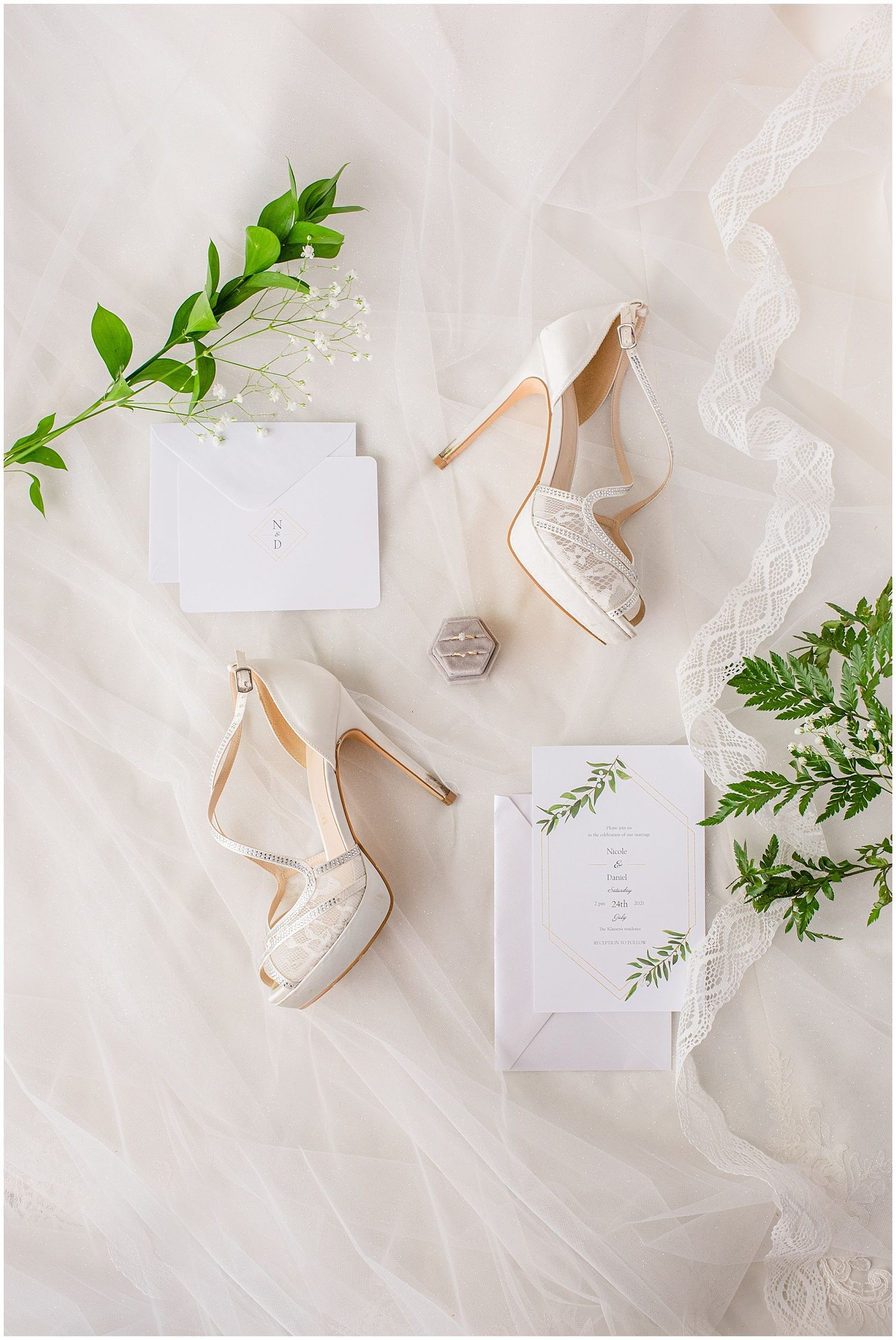 Elegant bridal flat lay with ivory lace heels, wedding invitation, ring box, and greenery on a white tulle veil.
