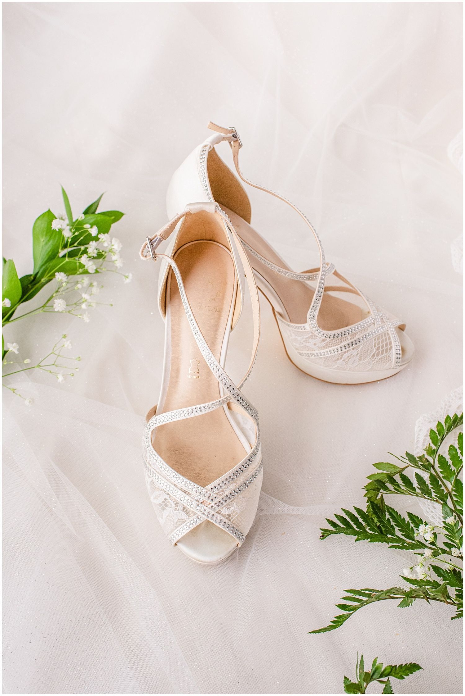 Elegant white bridal heels with crystal straps displayed on tulle fabric surrounded by greenery and baby's breath flowers.