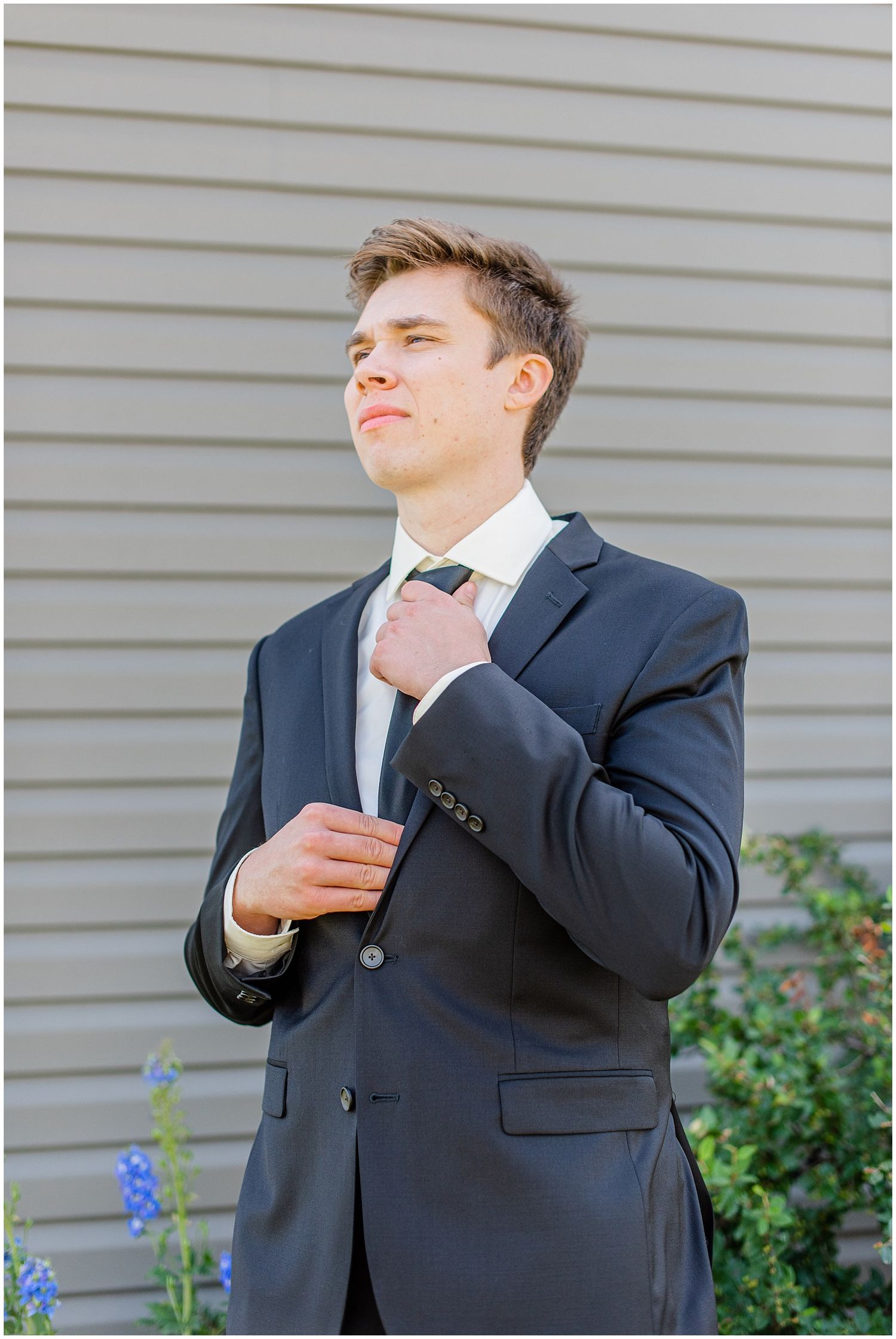 Young groom in dark suit adjusts his tie while standing outdoors before wedding ceremony.