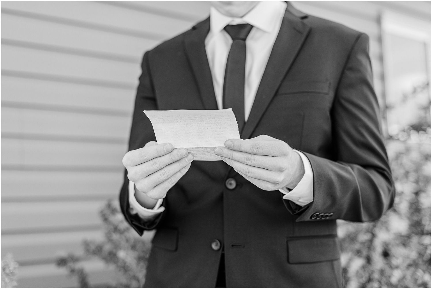 Black and white close-up of groom in dark suit reading a heartfelt letter before wedding ceremony.