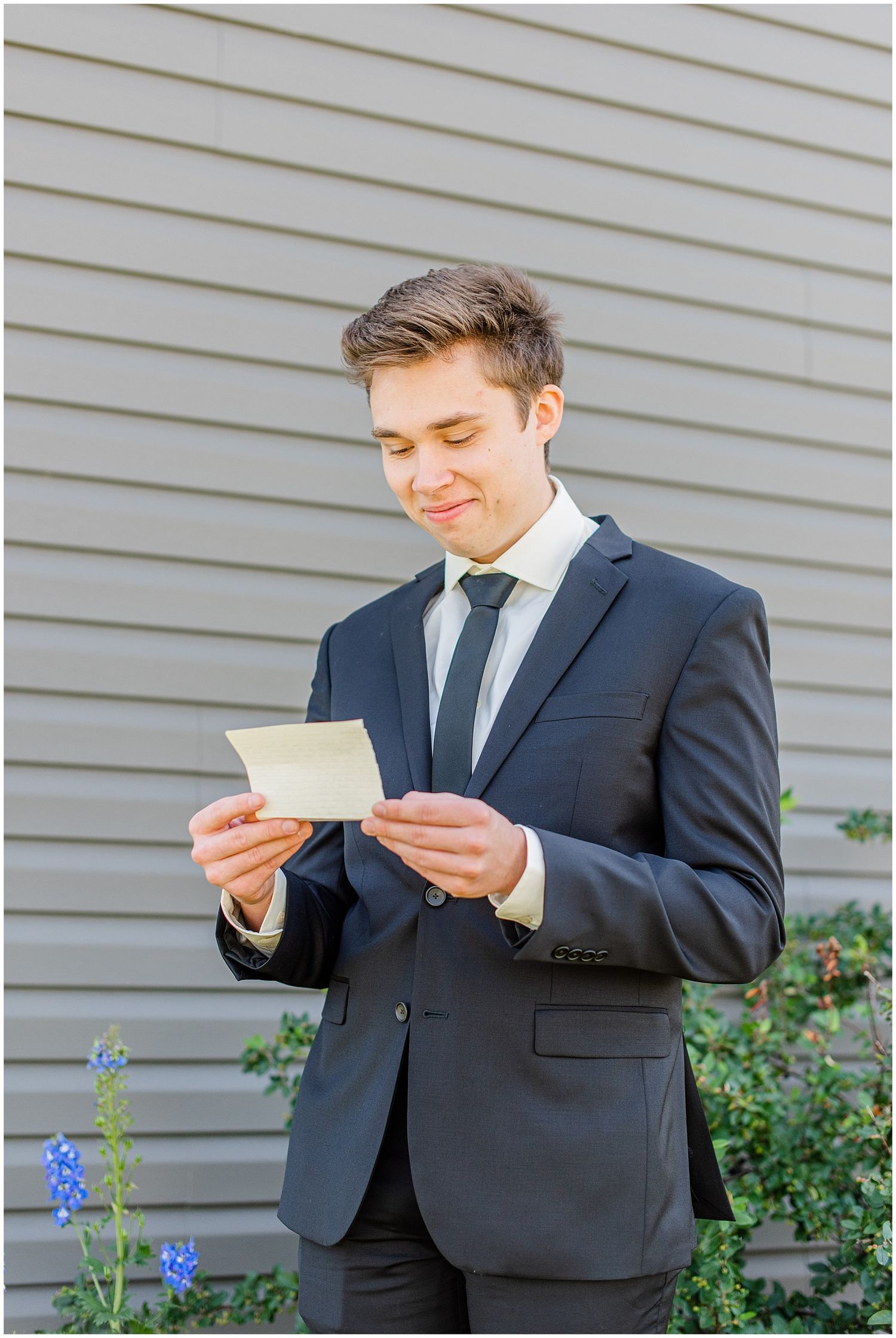 Handsome groom in dark suit reads a heartfelt letter while standing outside against a gray siding wall.