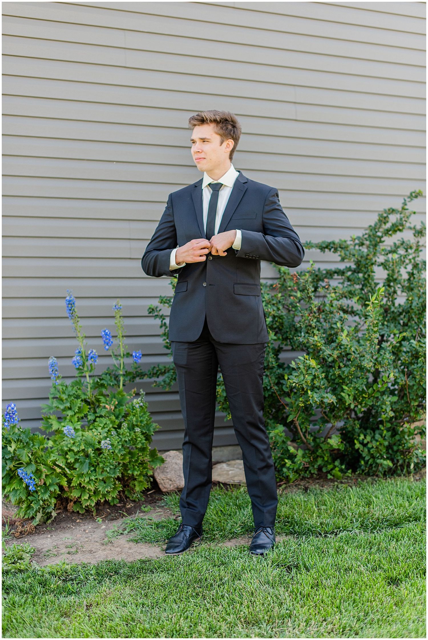 Dapper groom in black suit adjusting cufflinks outdoors beside gray building with flowers.