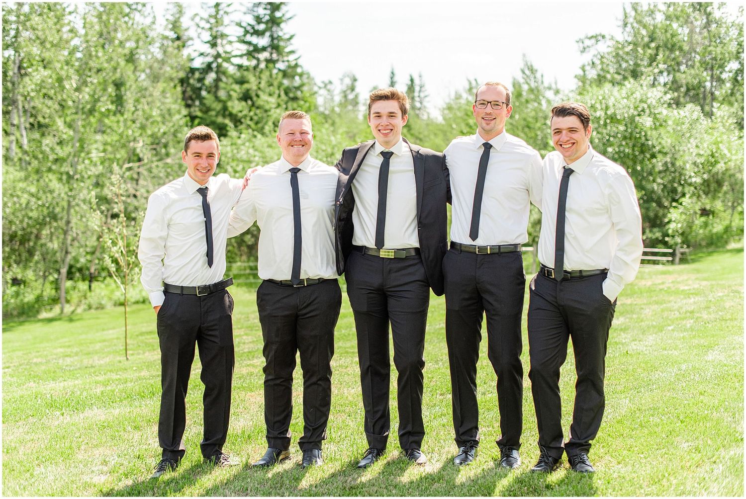 Five groomsmen in black suits and ties pose together outdoors on green grass.