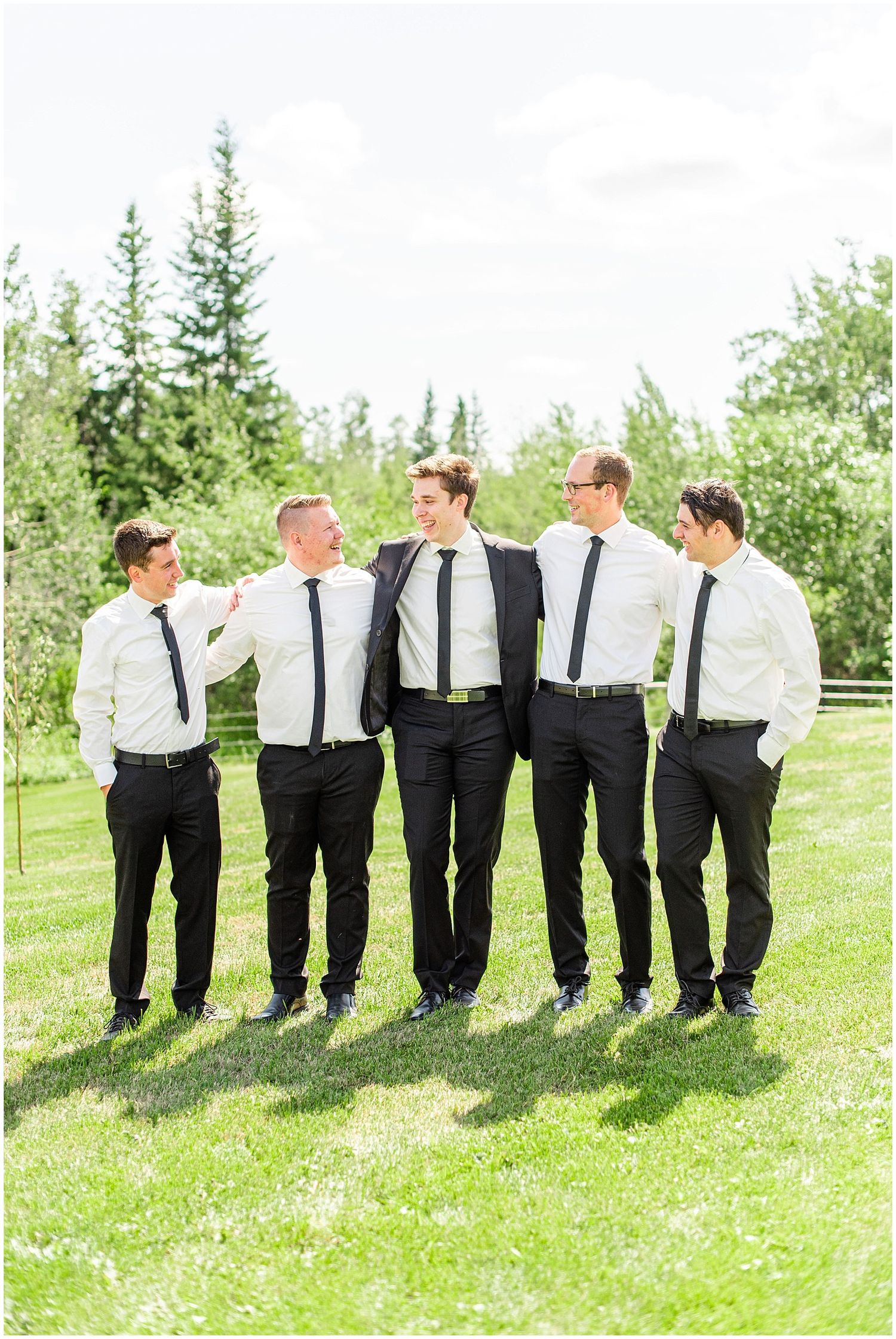 Groomsmen in black suits laugh and walk together on sunny green lawn at wedding.