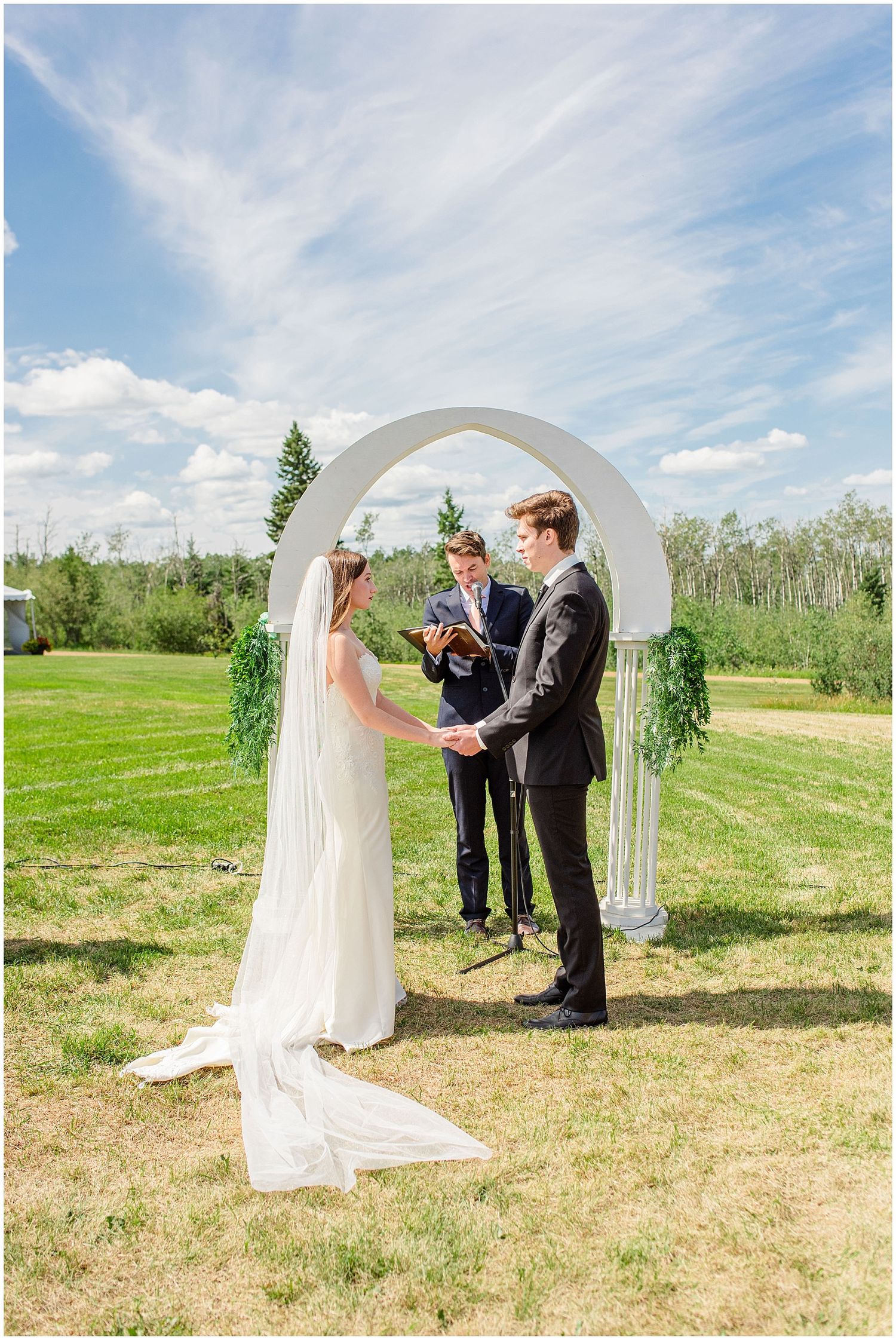 Bride and groom exchanging vows under white arch on sunny outdoor wedding ceremony.