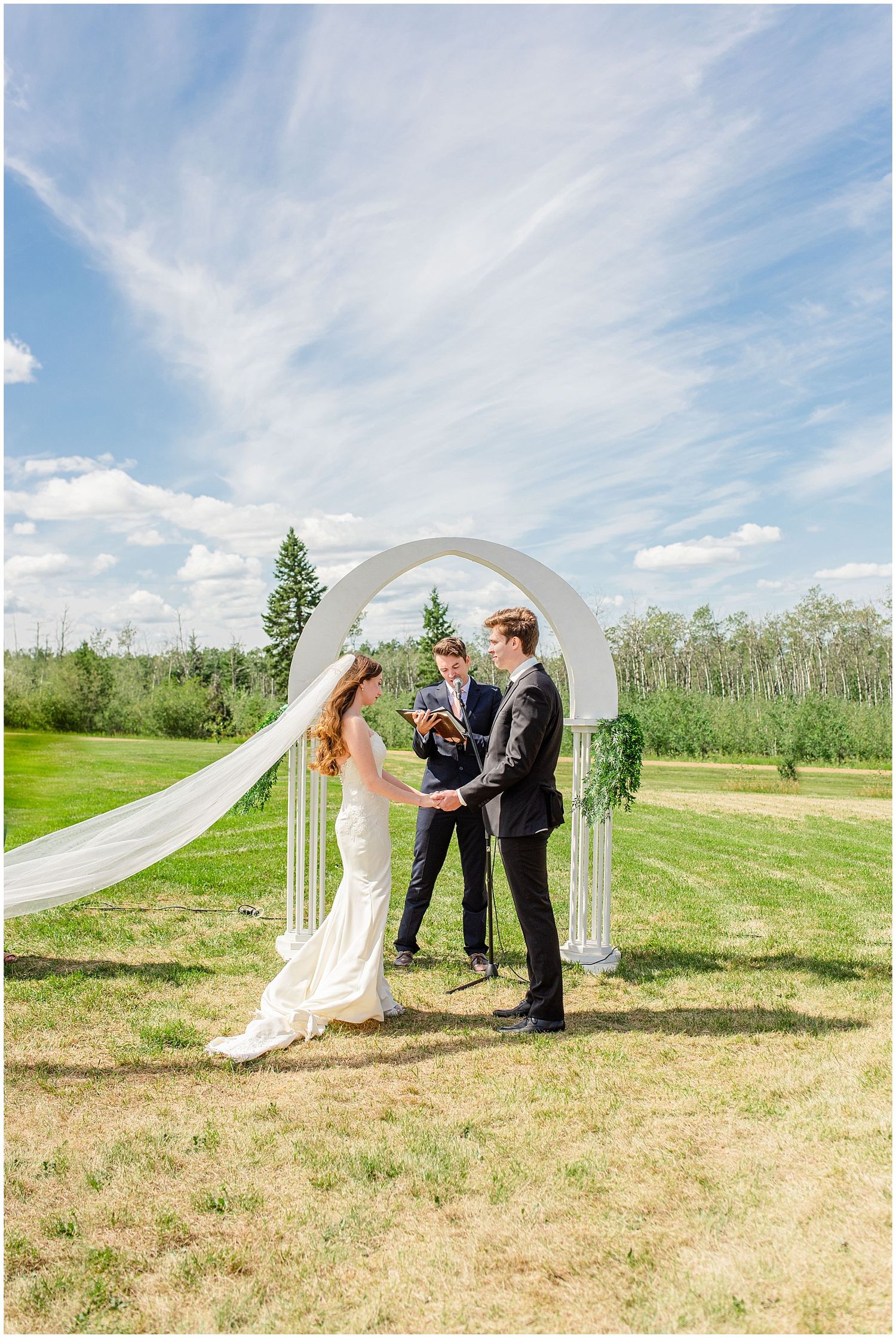 Bride and groom exchange vows outdoors under a white arch on a sunny day with a dramatic blue sky.