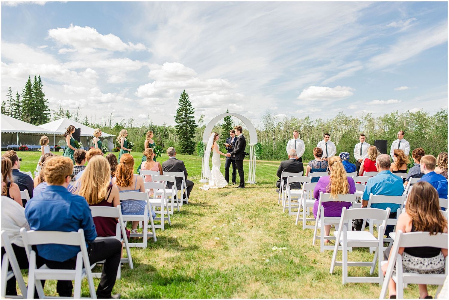 Guests seated in white chairs watch couple exchange vows at outdoor wedding ceremony.