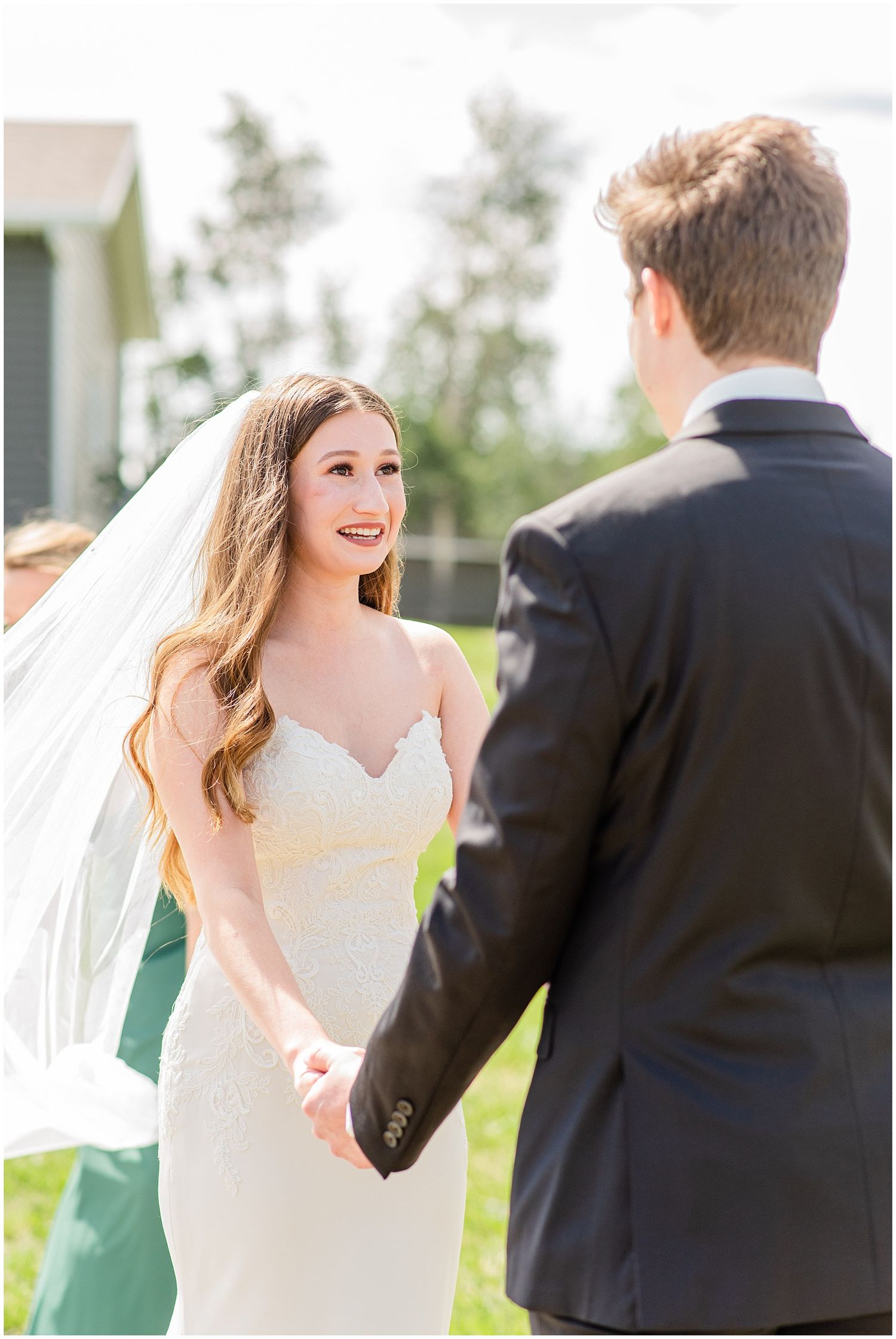 Smiling bride in lace gown holds hands with groom during outdoor wedding ceremony.