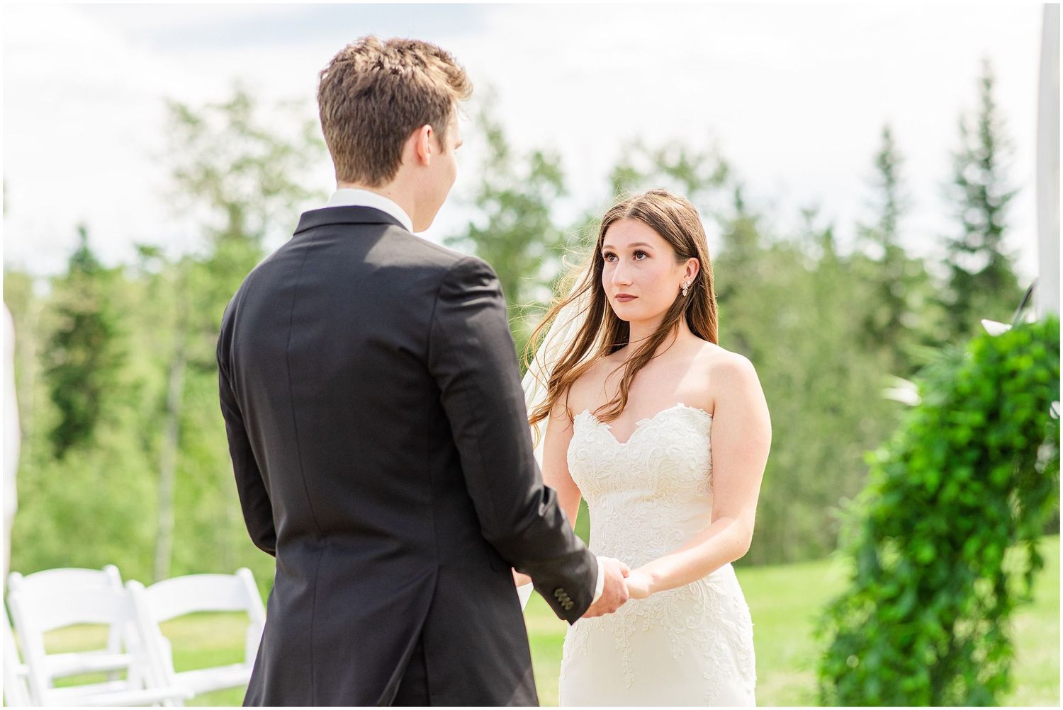 Bride and groom holding hands during outdoor wedding ceremony surrounded by lush green trees.