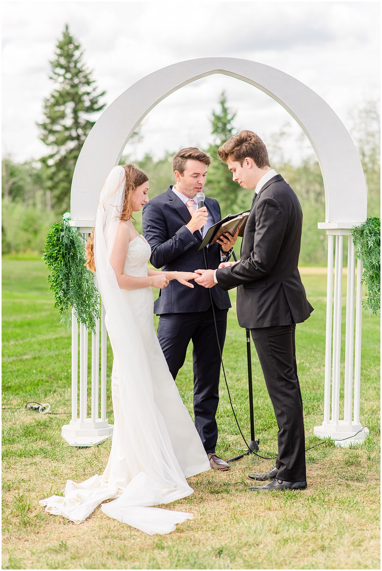 Bride and groom exchanging vows under white arch with greenery during outdoor wedding ceremony on lawn.