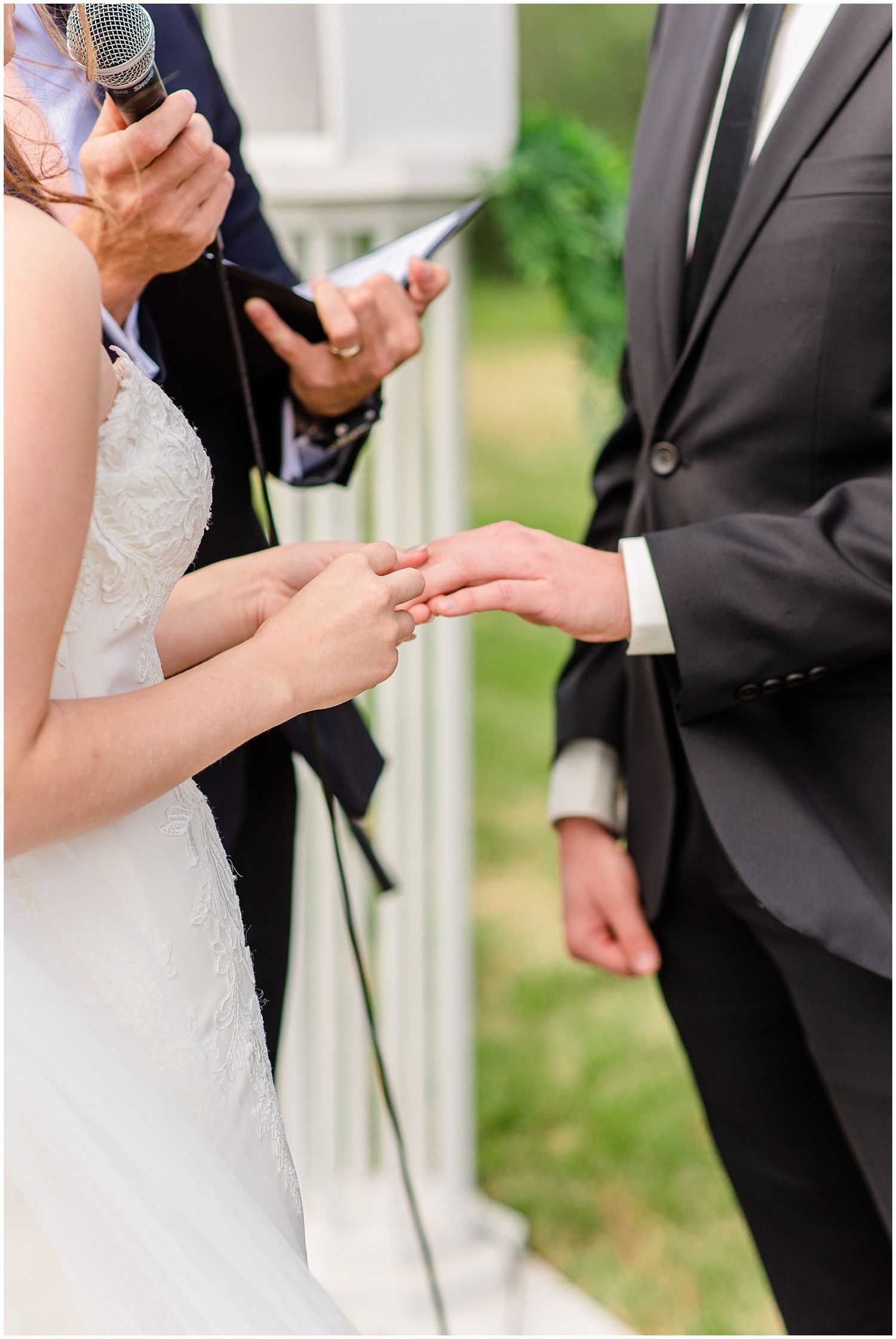 Bride places wedding ring on grooms finger during outdoor wedding ceremony officiated by minister.