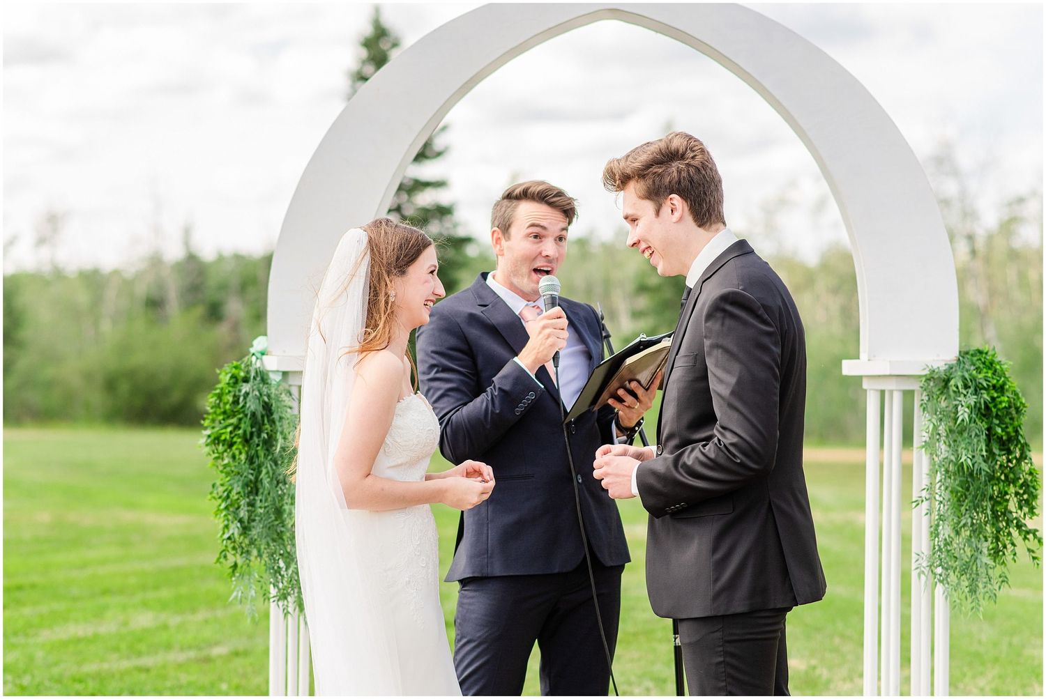 Officiant laughs with bride and groom during joyful outdoor wedding ceremony at white arch.
