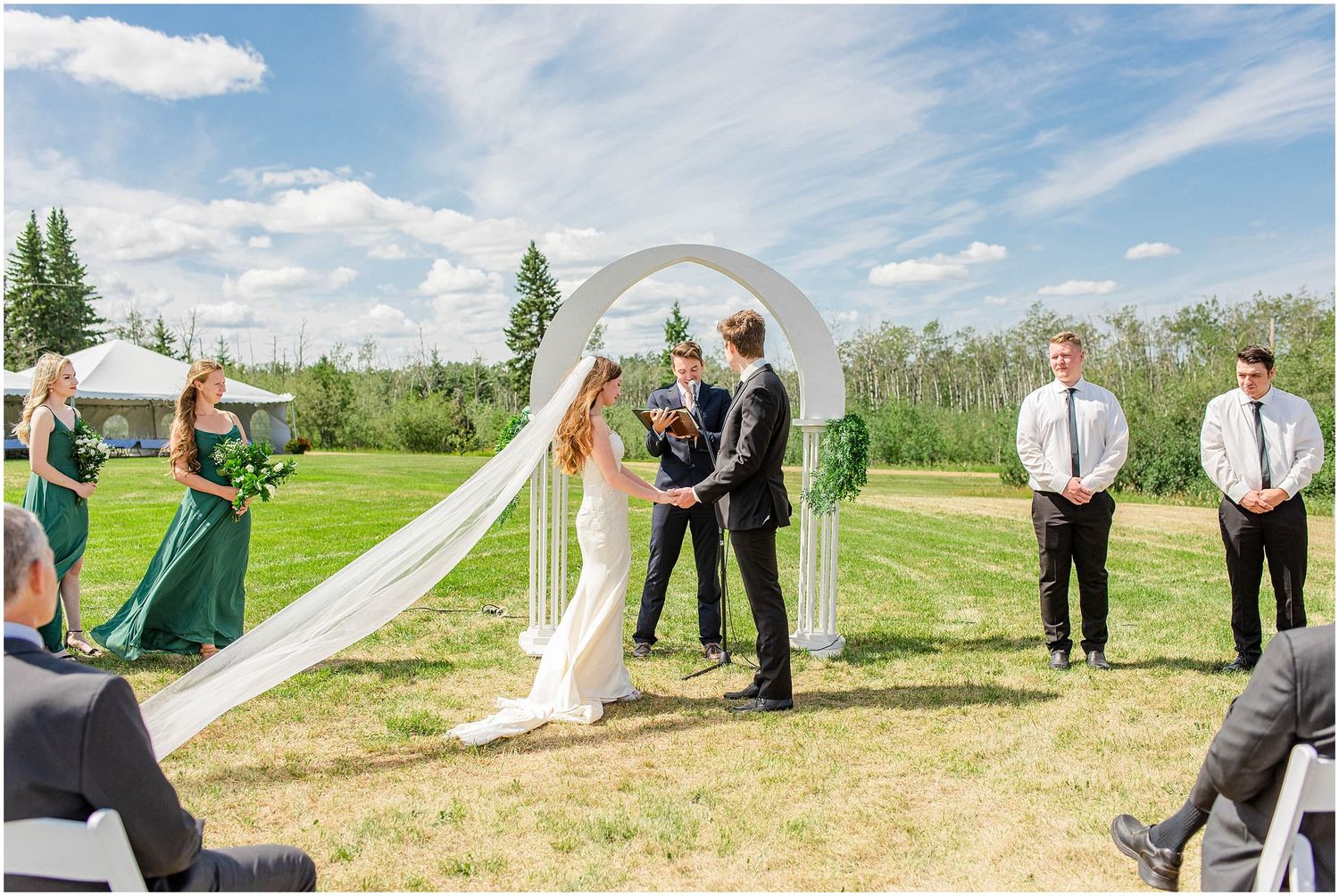 Outdoor wedding ceremony under white arch with bride and groom exchanging vows on green lawn.
