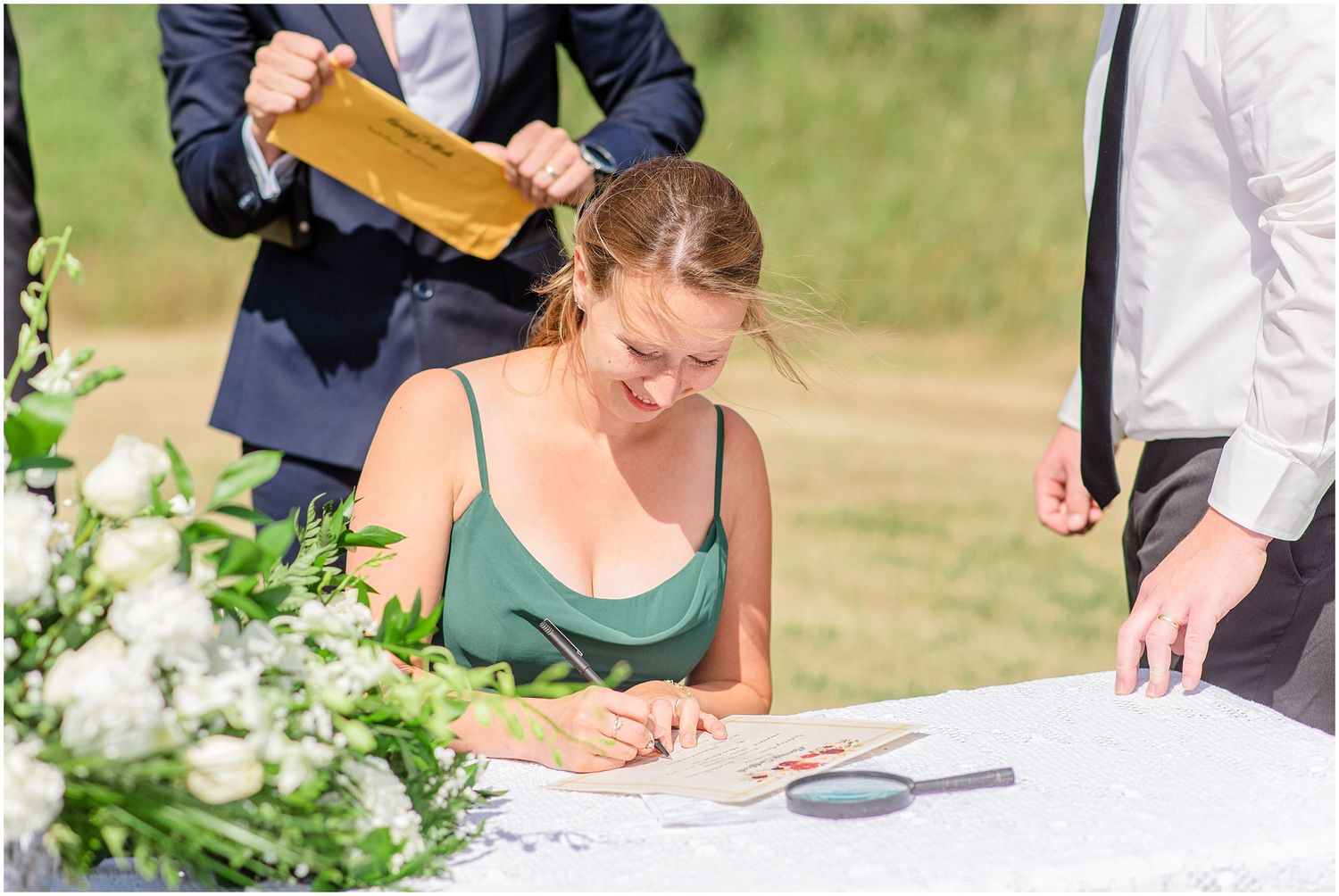 Wedding guest in green dress signs official marriage documents at an outdoor ceremony table.