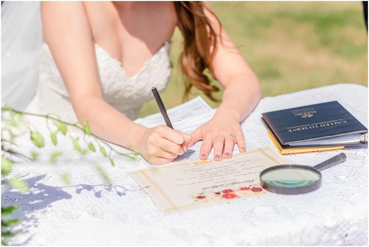 Bride signing wedding certificate at outdoor ceremony table with magnifying glass and passport nearby.