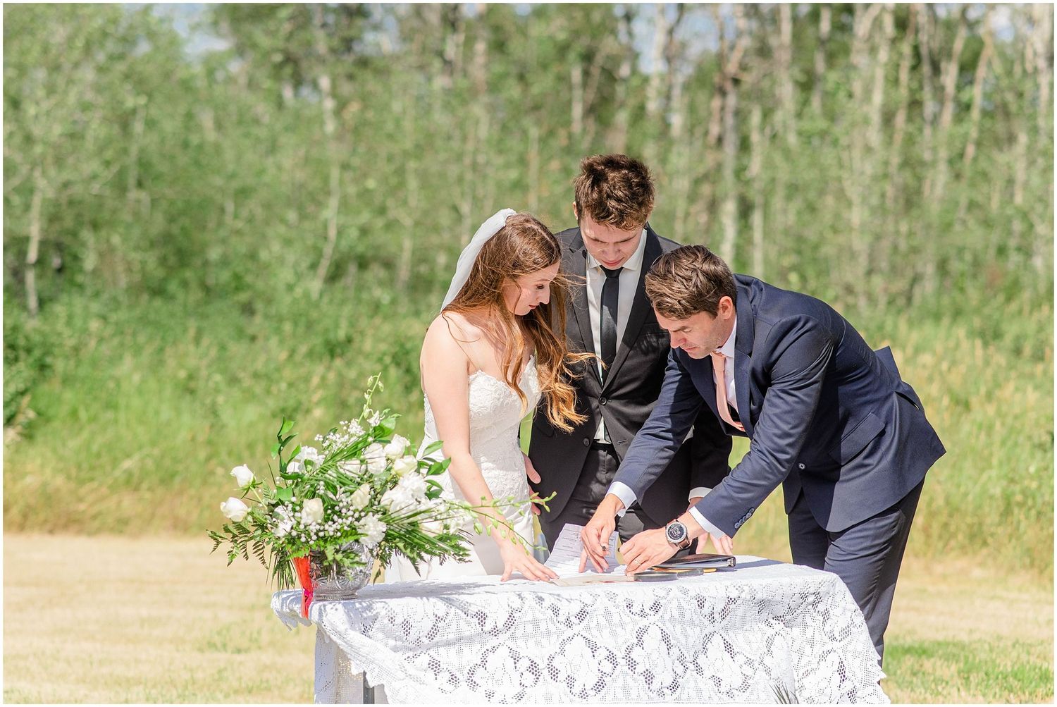 Bride, groom, and officiant sign wedding documents at lace-covered table outdoors.