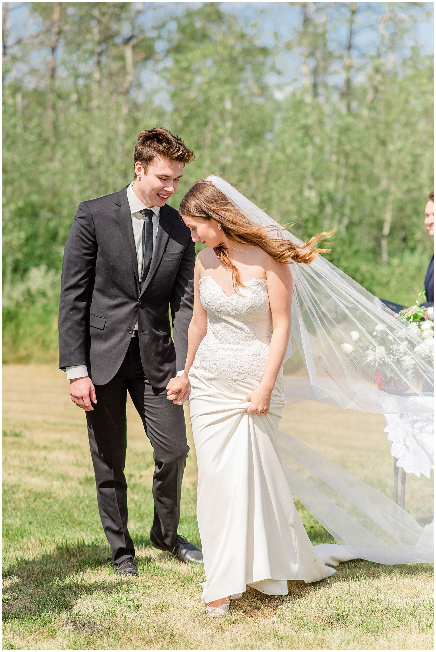Newlyweds walk hand in hand on green lawn, bride's veil flowing in the breeze.