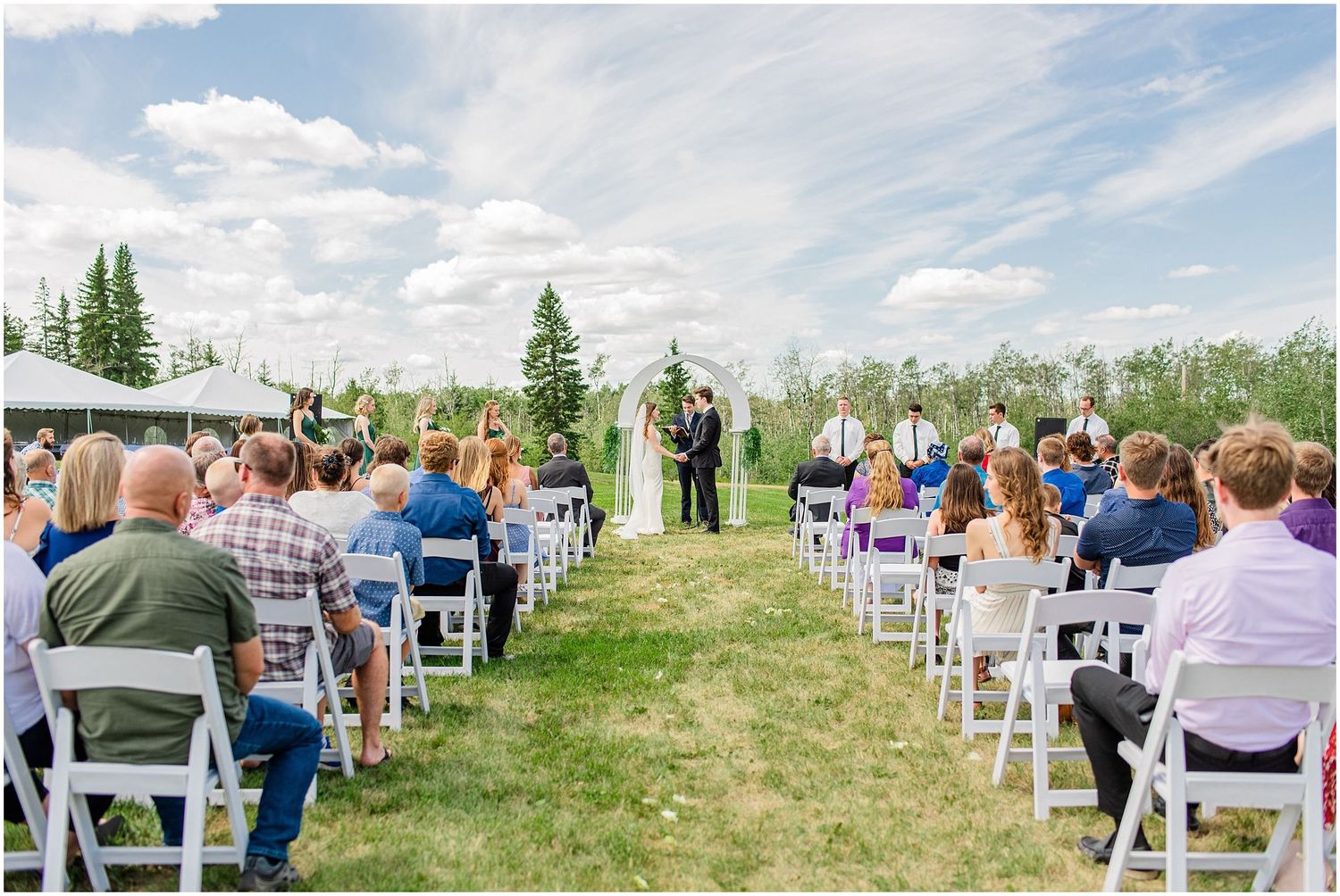 Guests seated in white chairs watch an outdoor wedding ceremony on a sunny day with lush green trees.