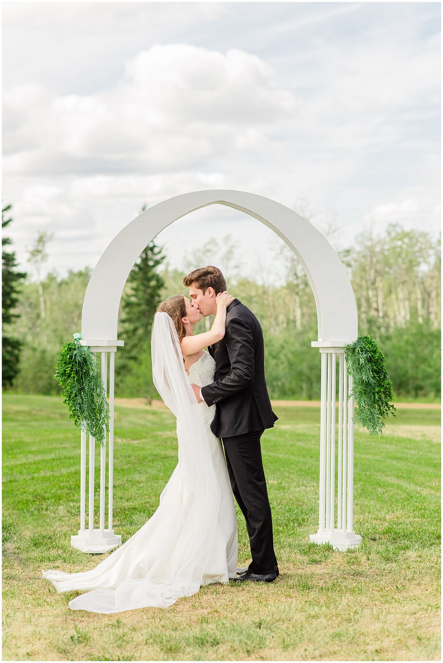 Bride and groom share first kiss under white arch with greenery on lush lawn.