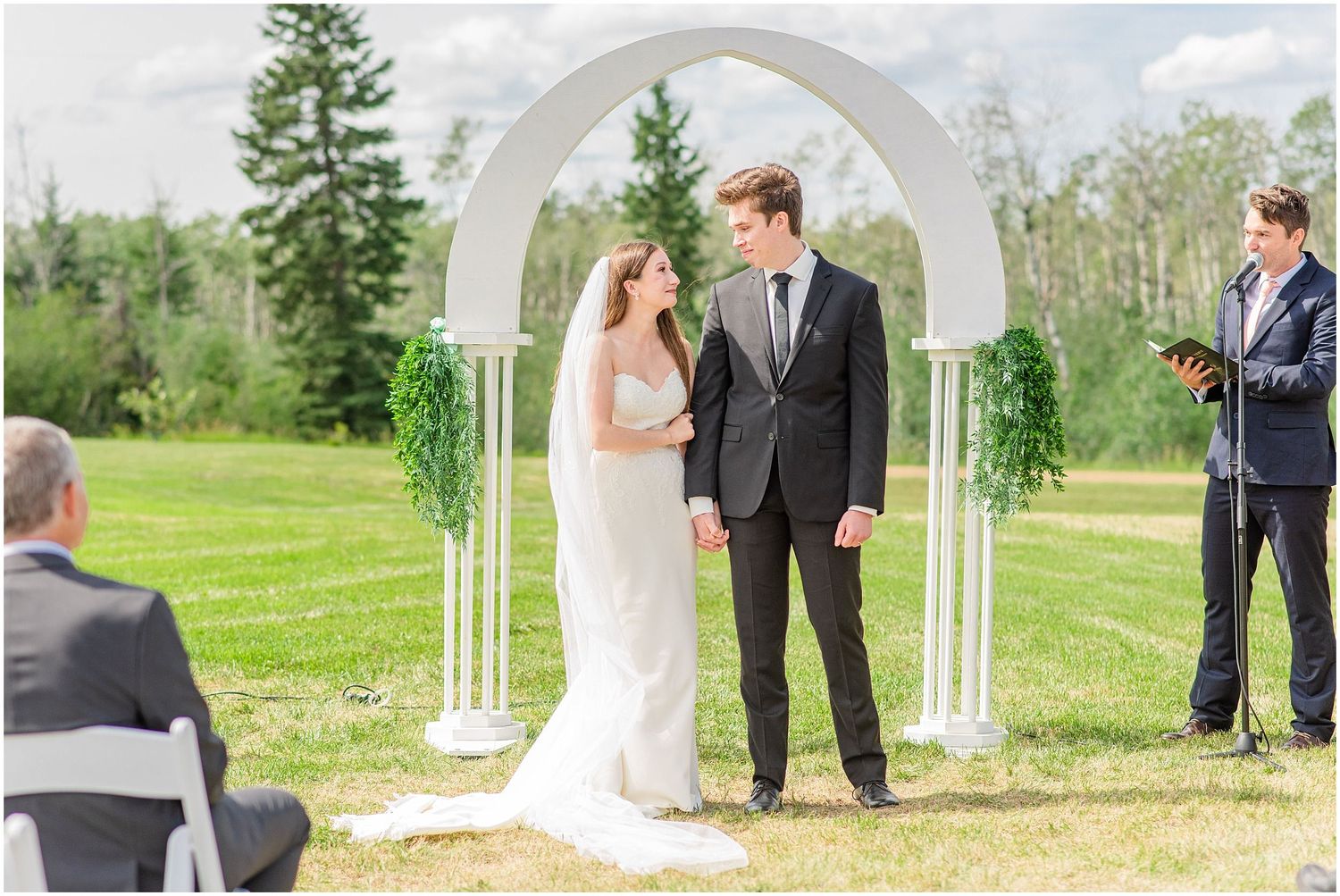 Bride and groom share a loving glance under a white arch during their outdoor wedding ceremony.