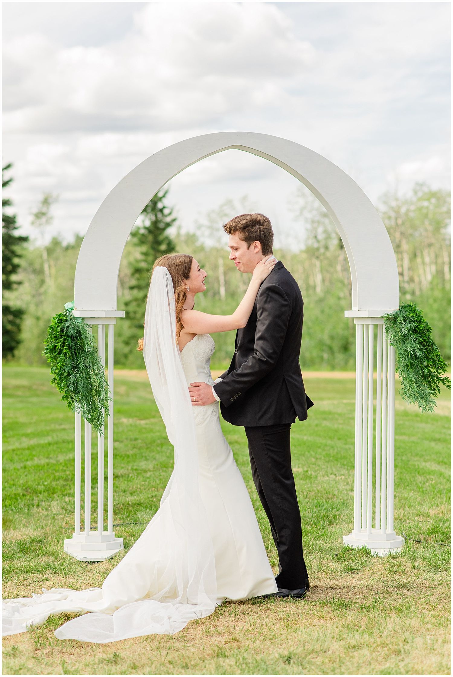 Bride and groom embrace under white arch with greenery at outdoor wedding ceremony.