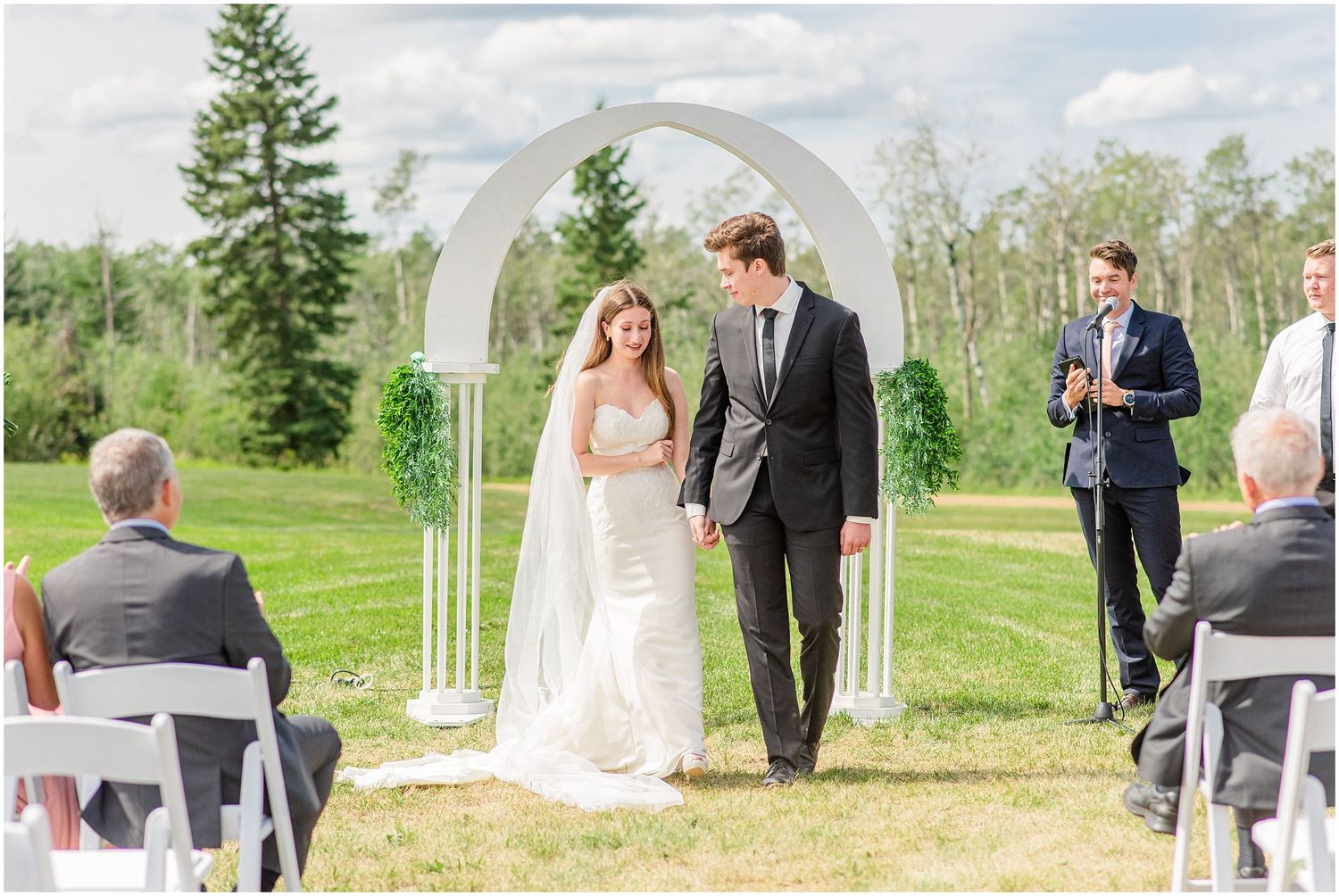 Bride and groom walk hand in hand under a white arch at an outdoor wedding ceremony on a sunny day.