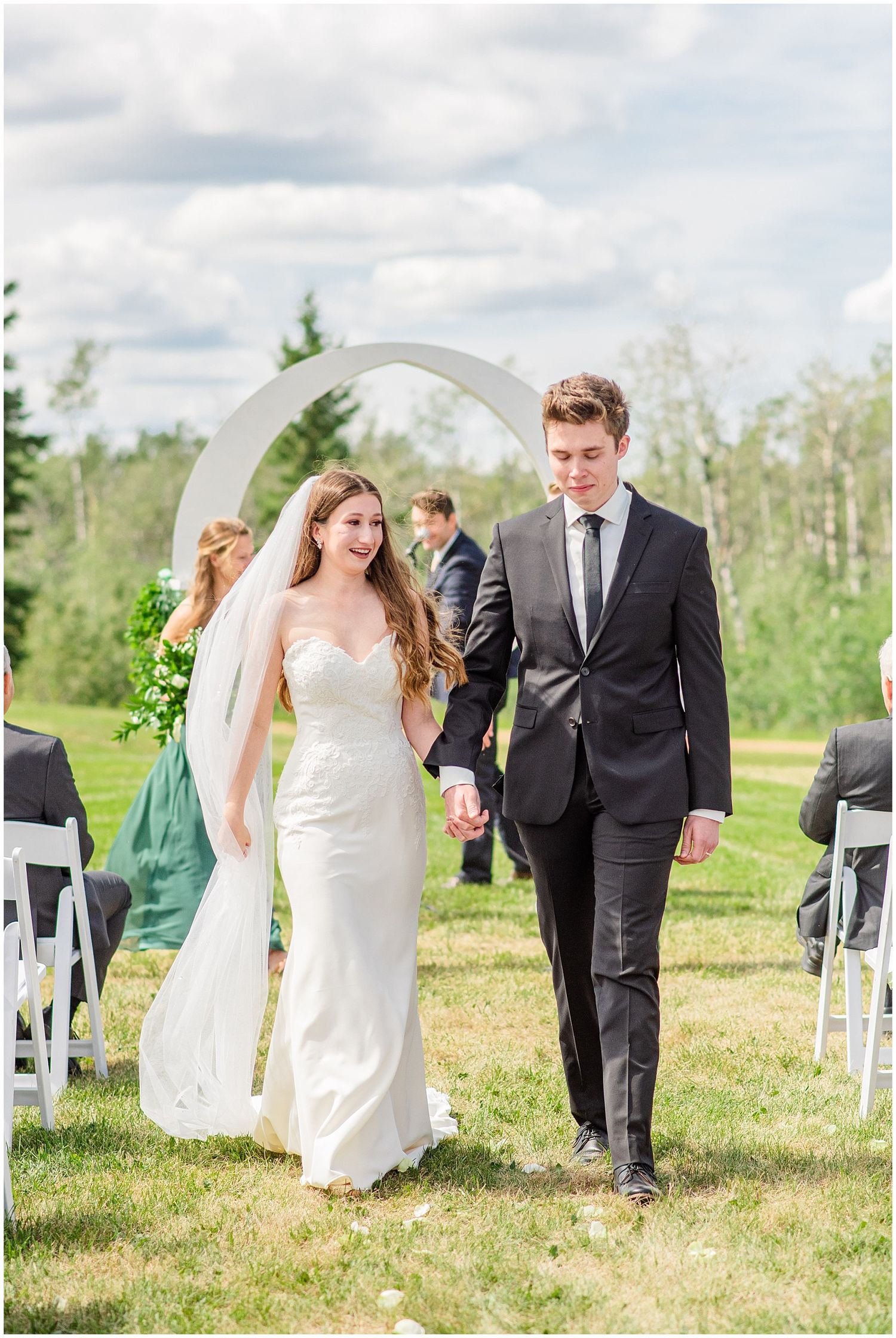 Newlyweds smiling and walking back down the aisle under a white arch after outdoor wedding ceremony.