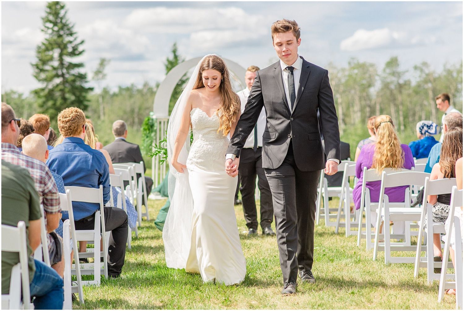 Newlywed bride and groom walk hand in hand back down the aisle after outdoor wedding ceremony.