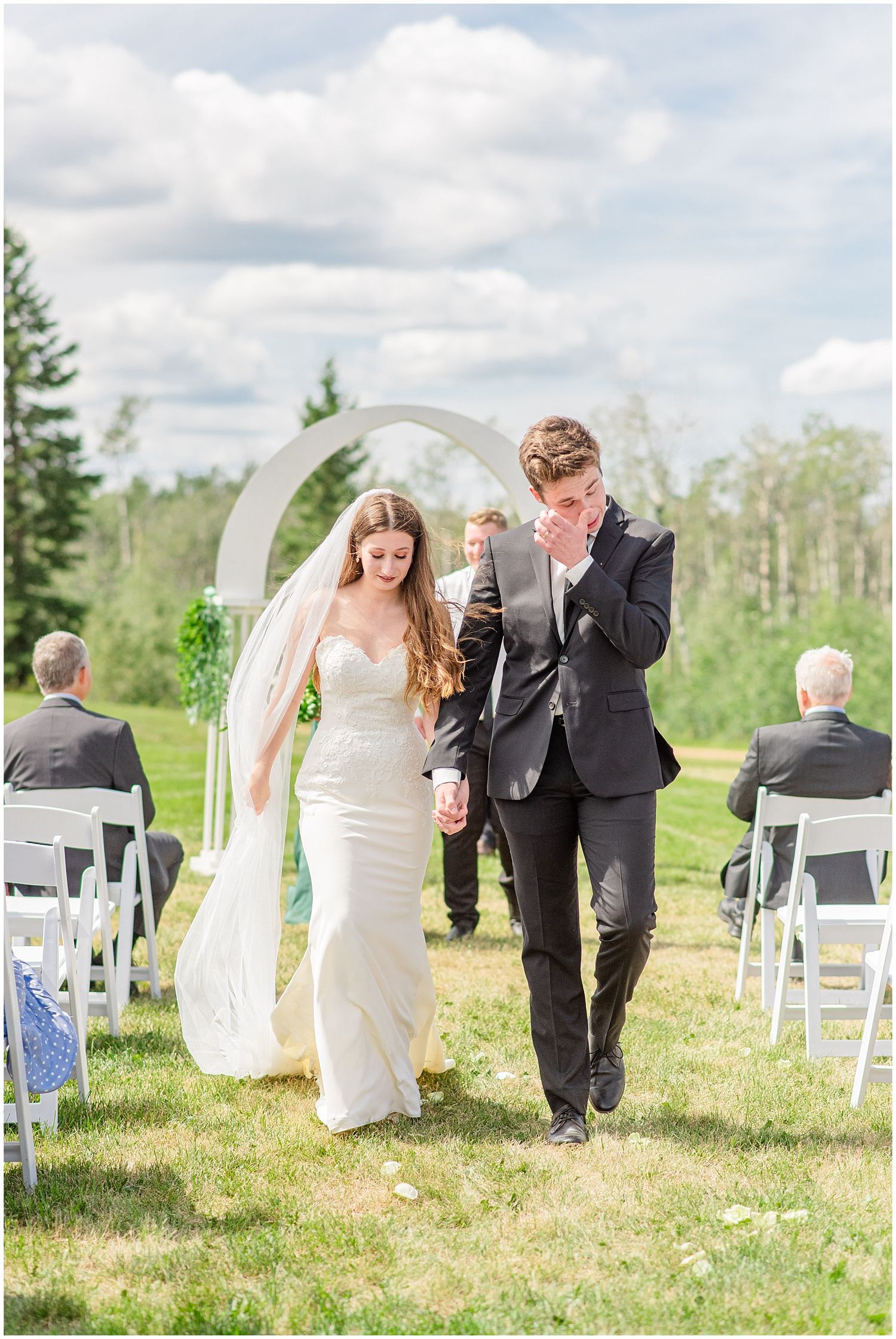 Emotional groom wipes tears as he walks with his bride down the aisle after their outdoor ceremony.