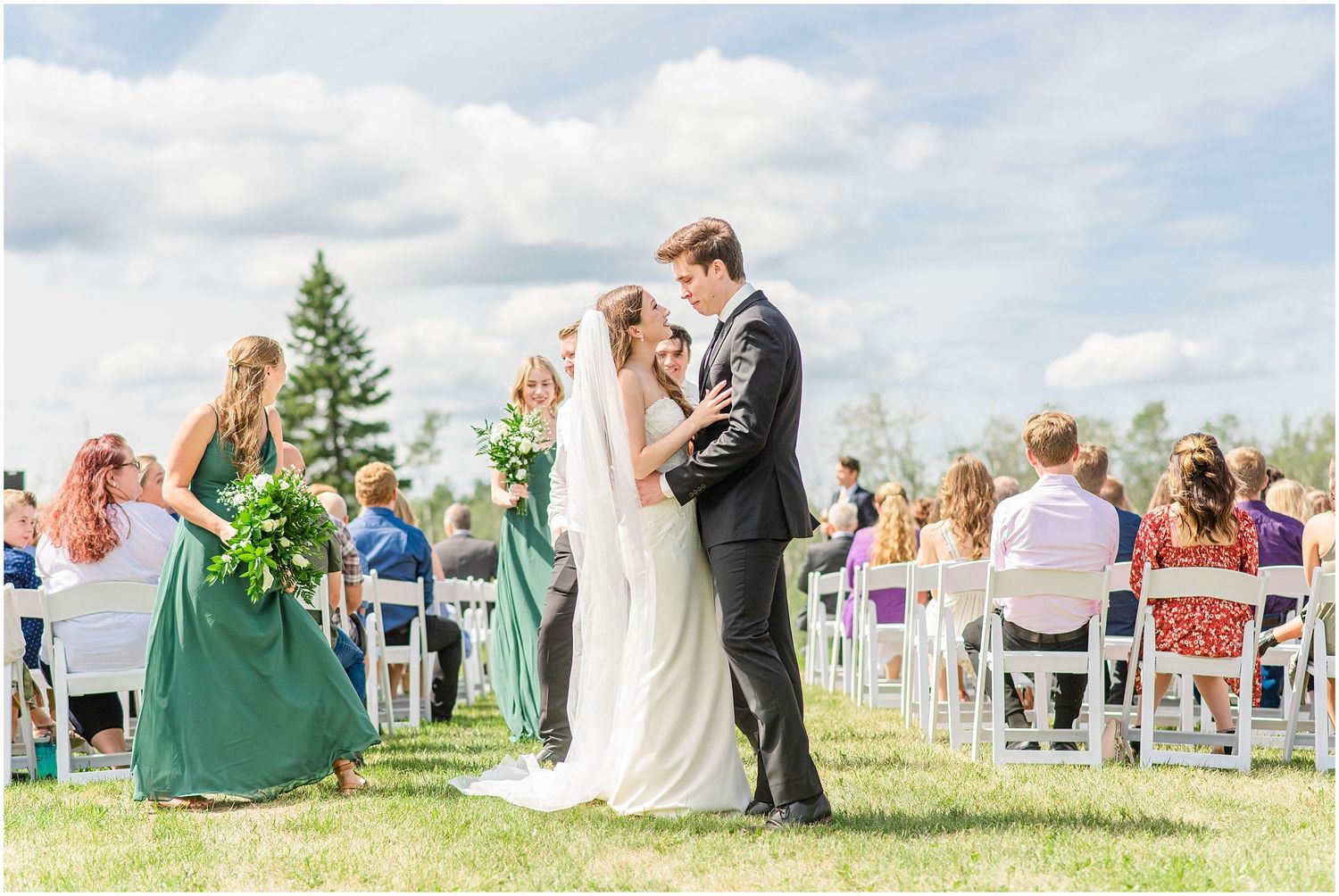 Newlyweds sharing first kiss at outdoor ceremony with green-dressed bridesmaids nearby.