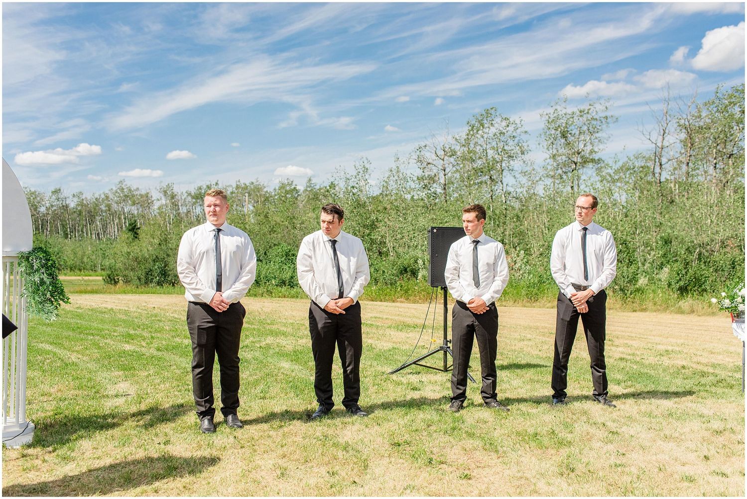Four groomsmen in white shirts and black ties stand in a row at outdoor wedding ceremony.