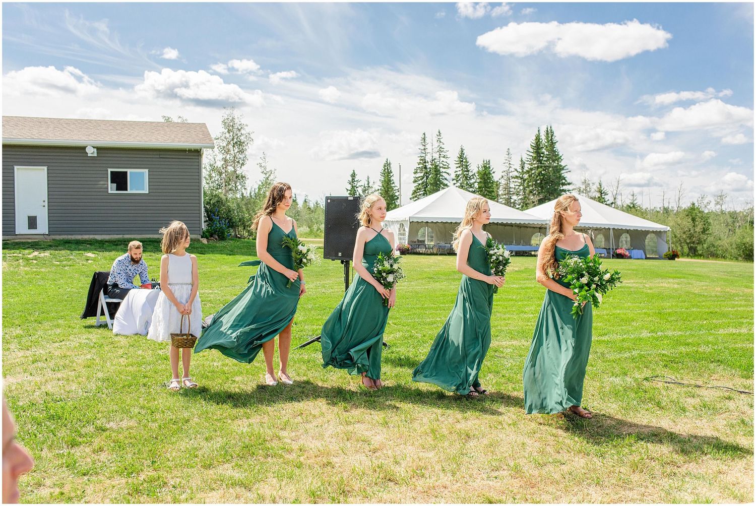 Bridesmaids in green dresses walk across lawn at outdoor wedding ceremony.