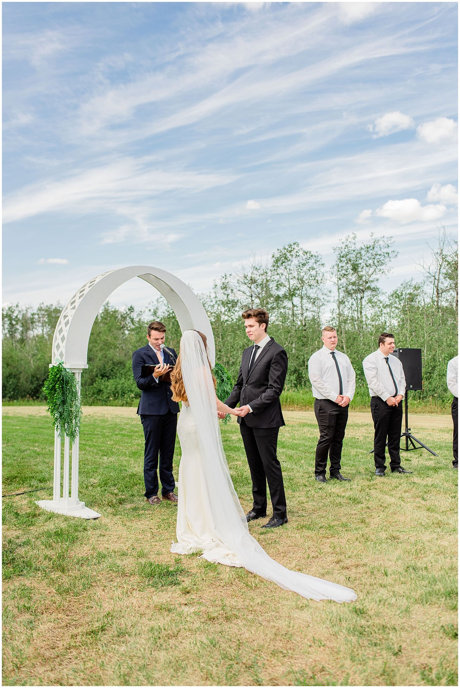 Bride and groom exchange vows under white arch at outdoor wedding ceremony.