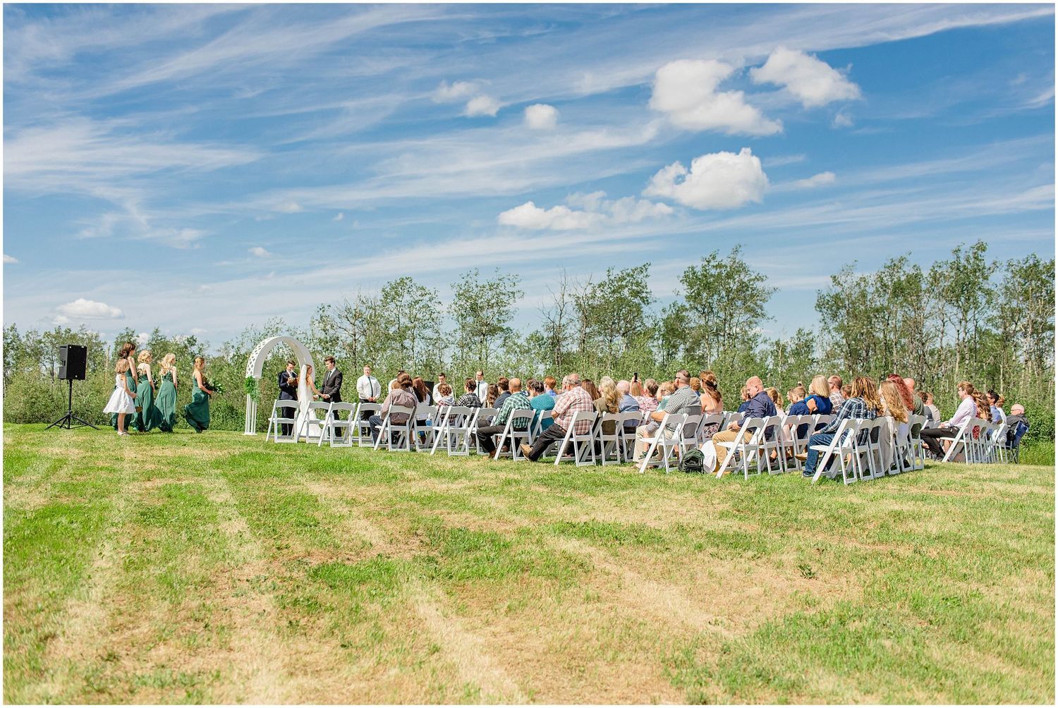 Outdoor wedding ceremony with guests seated in white chairs on a green lawn under a bright blue sky.