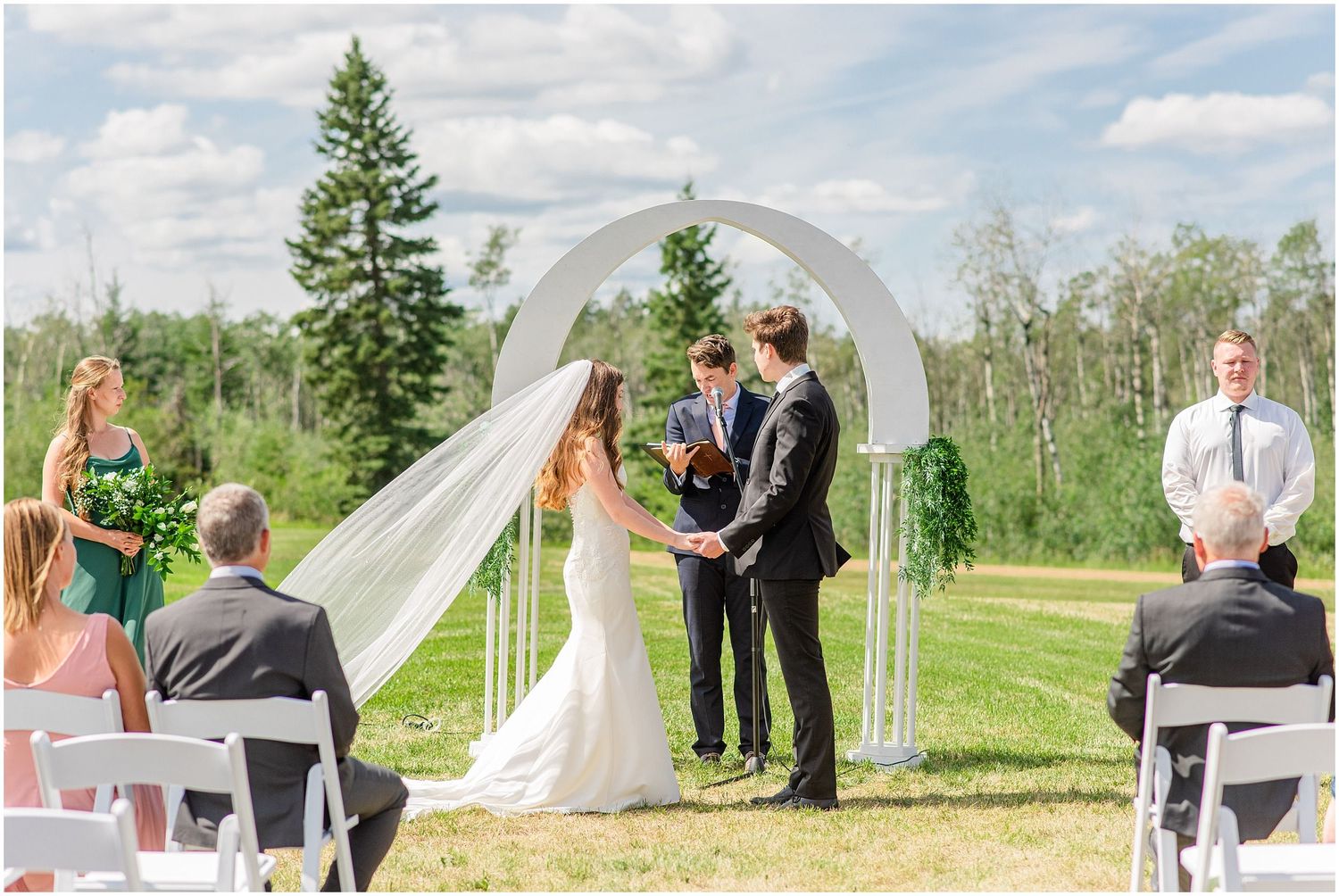 Bride and groom exchange vows at an outdoor wedding ceremony under a floral arch on a sunny day.