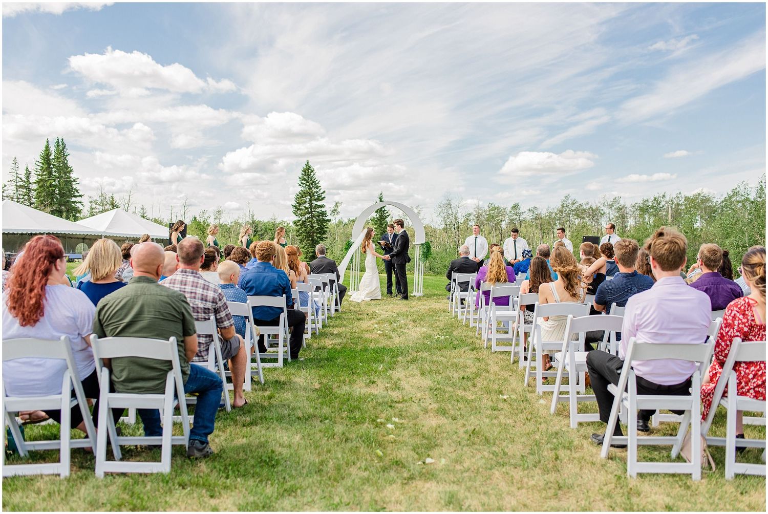 Guests seated in white chairs watch bride and groom exchange vows at outdoor summer wedding ceremony.