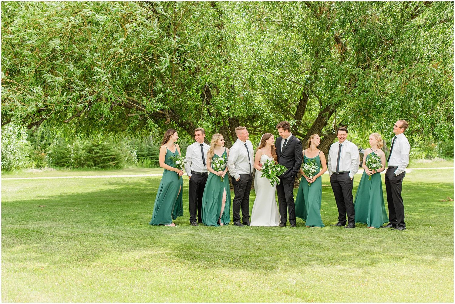Wedding party in green dresses and black suits posing outdoors under a large tree on a sunny day.