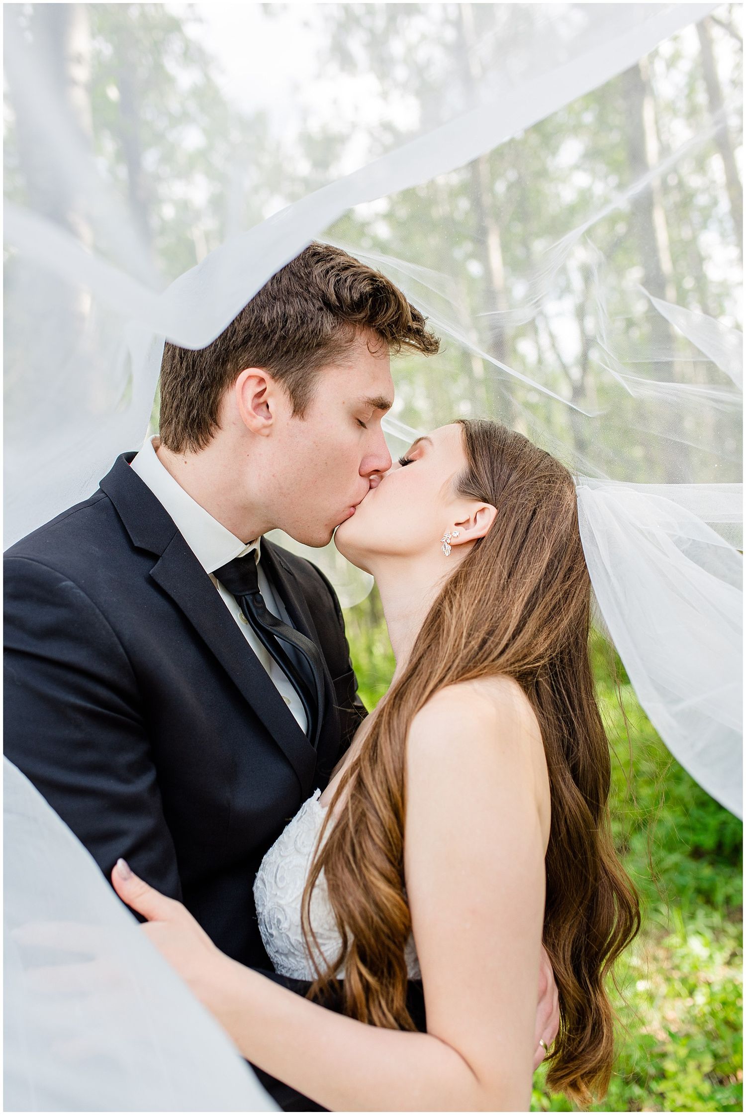 Bride and groom sharing a romantic kiss beneath a flowing white veil in a lush green forest setting.