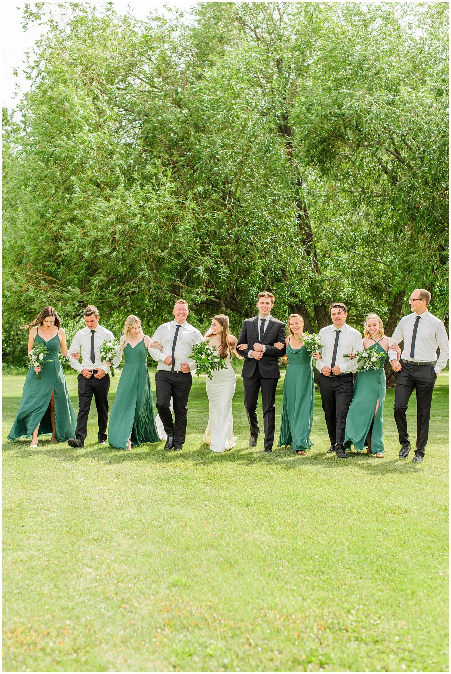 Wedding party walks together on green lawn, bridesmaids in emerald dresses and groomsmen in black suits.