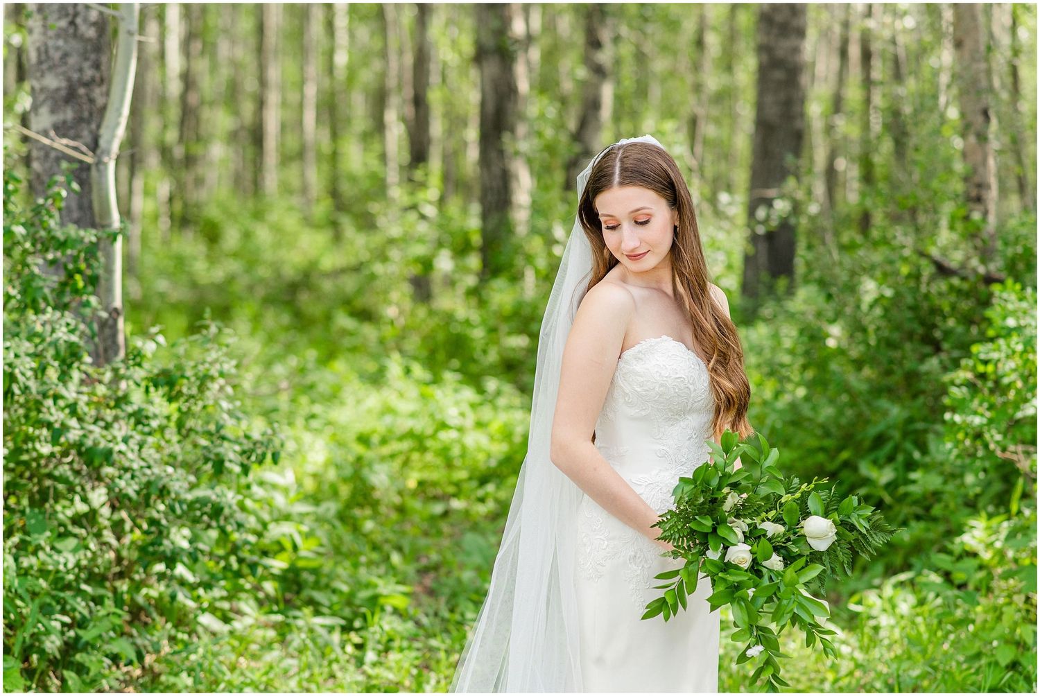Bride in a strapless gown and long veil holds a bouquet while standing among lush green forest foliage.